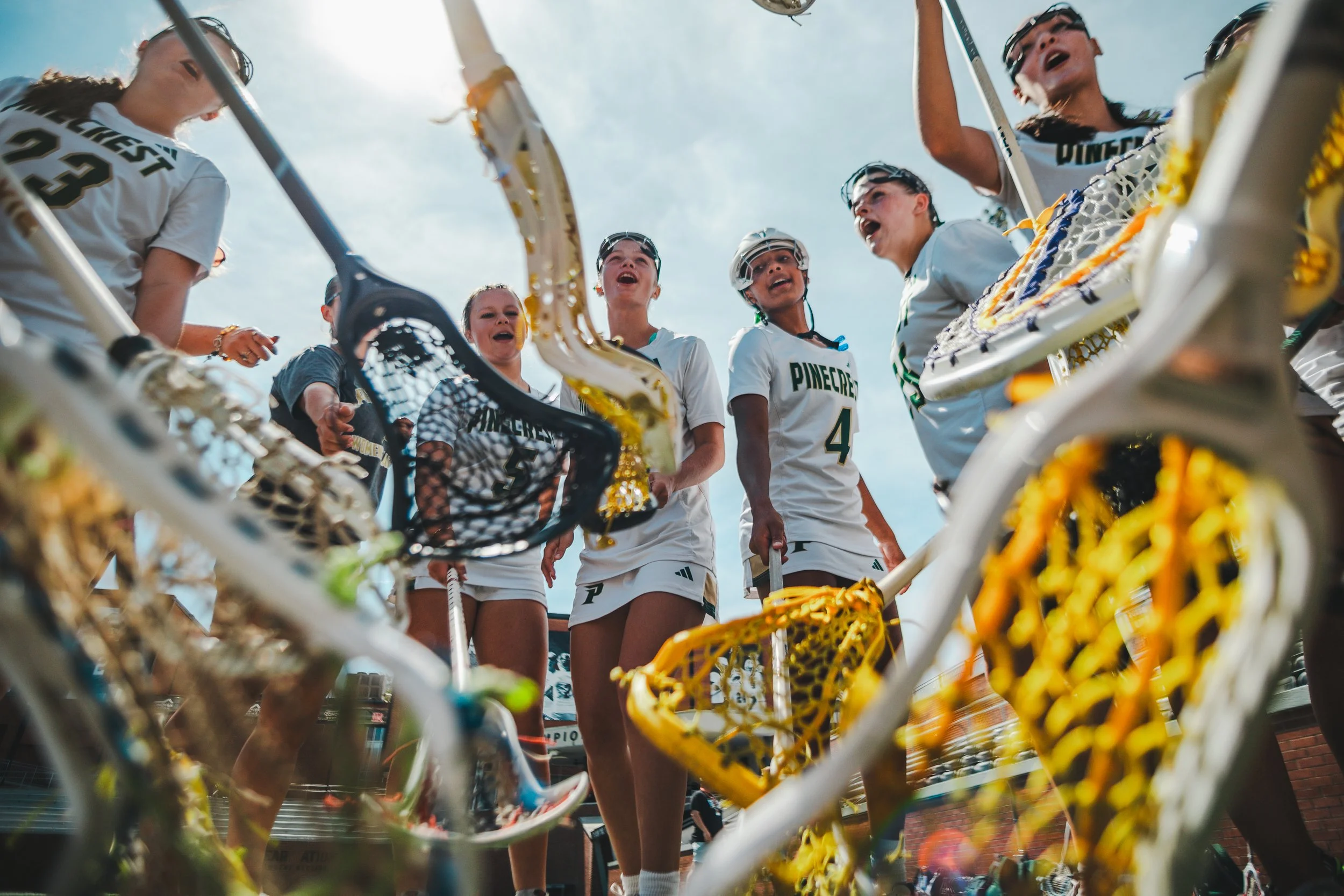 A group of female lacrosse players in white uniforms standing together on the field, viewed from below with lacrosse sticks in the foreground.