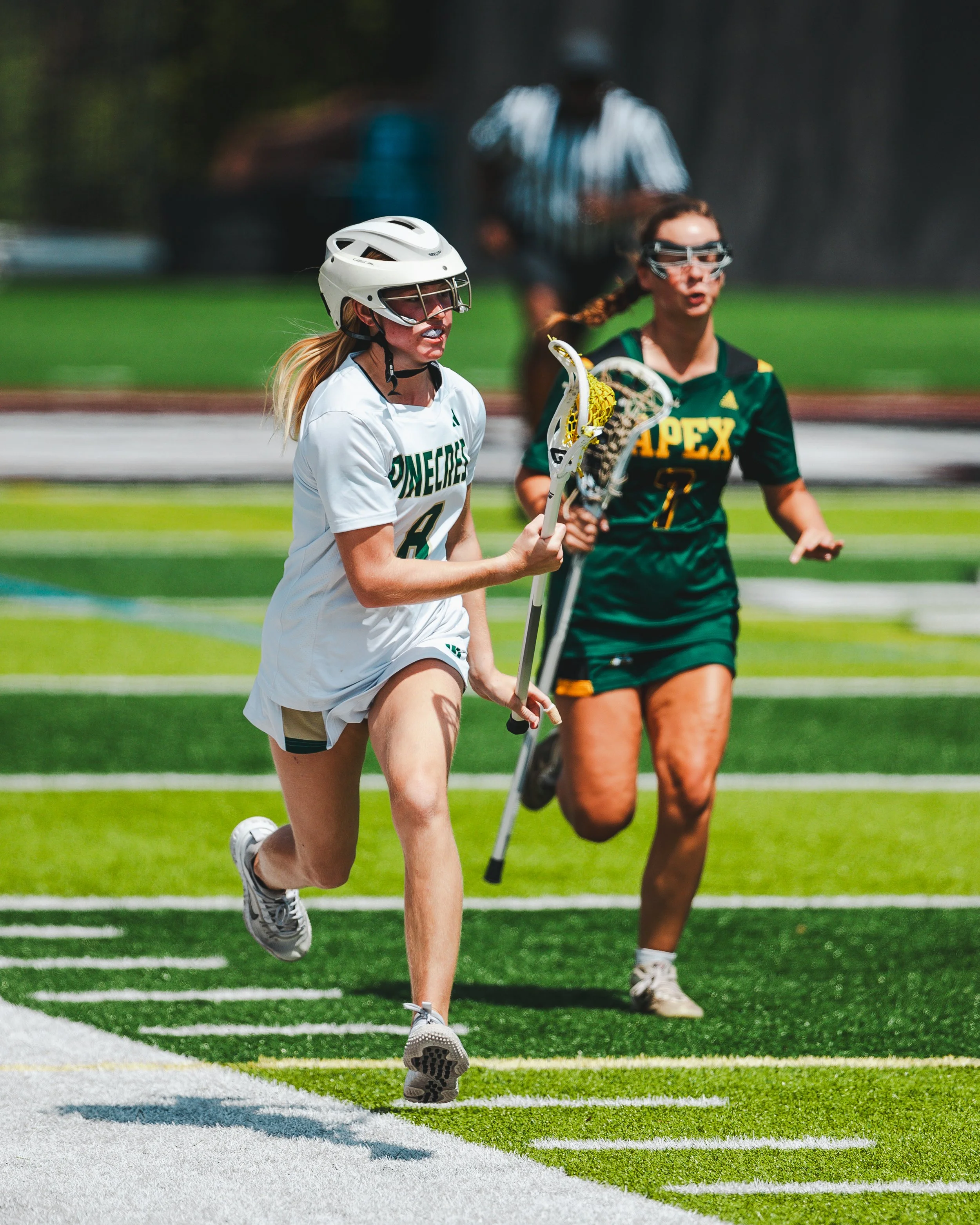 A girl playing lacrosse wearing safety goggles and a white helmet running on a sports field with a lacrosse stick in hand, alongside another girl in a green sports jersey and goggles.