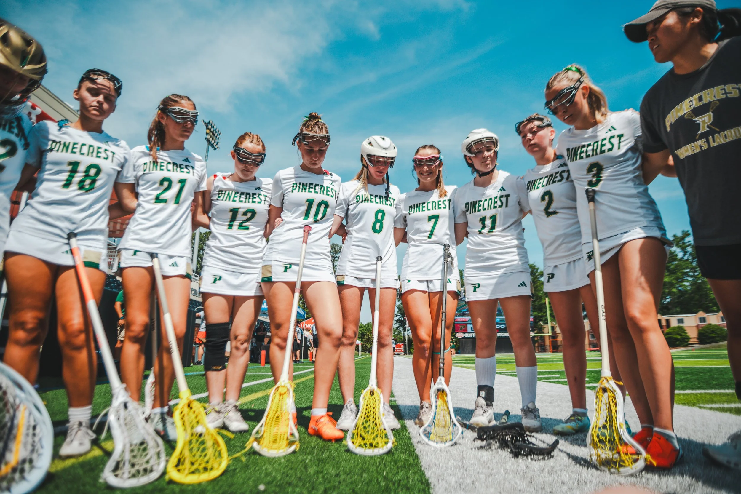 A group of female lacrosse players standing in a team huddle on a sports field, wearing white jerseys with green text, holding lacrosse sticks, with a blue sky and stadium lights in the background.