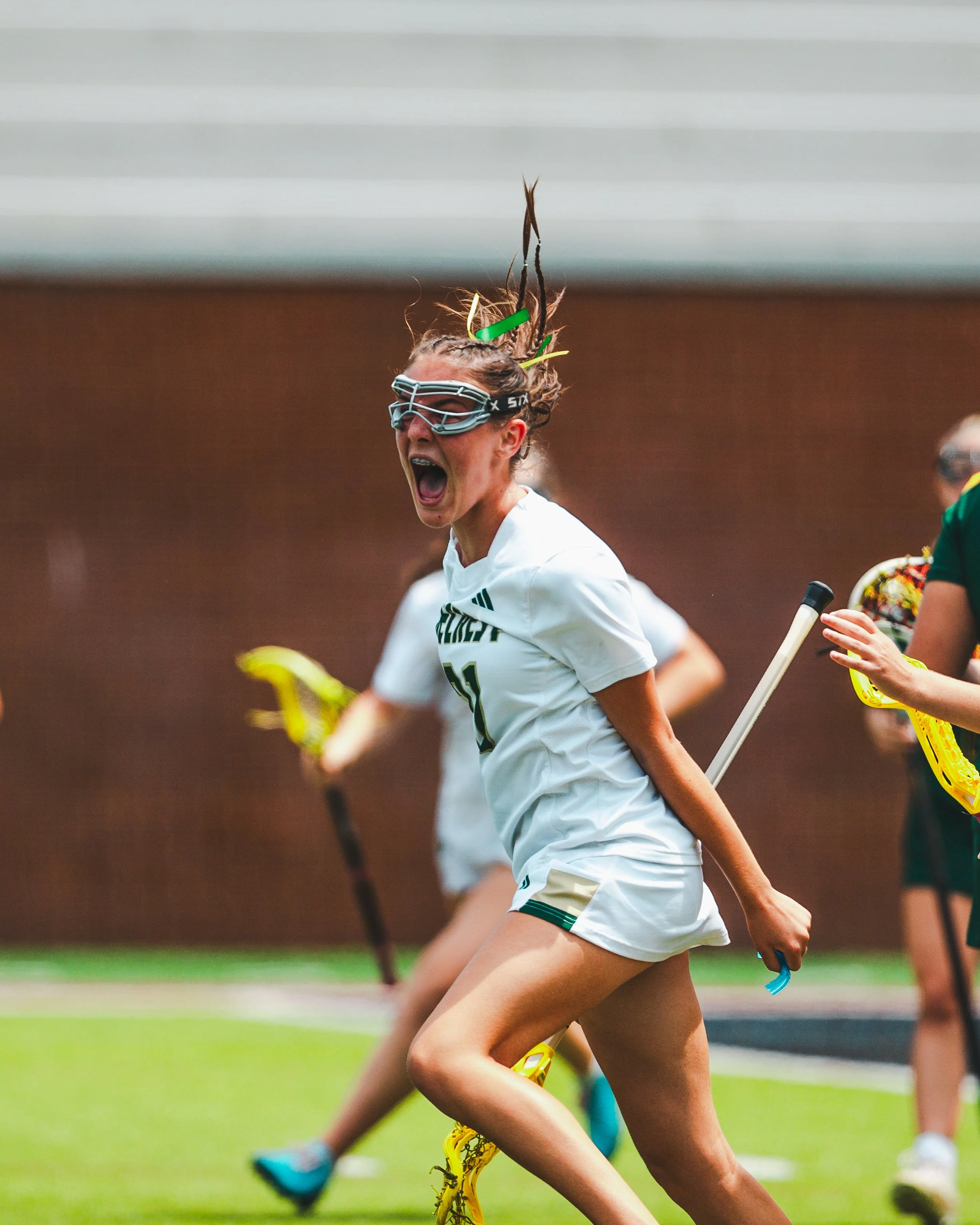 Close-up of a female athlete wearing protective eyewear, running on a field during a sports event, with other athletes in the background.