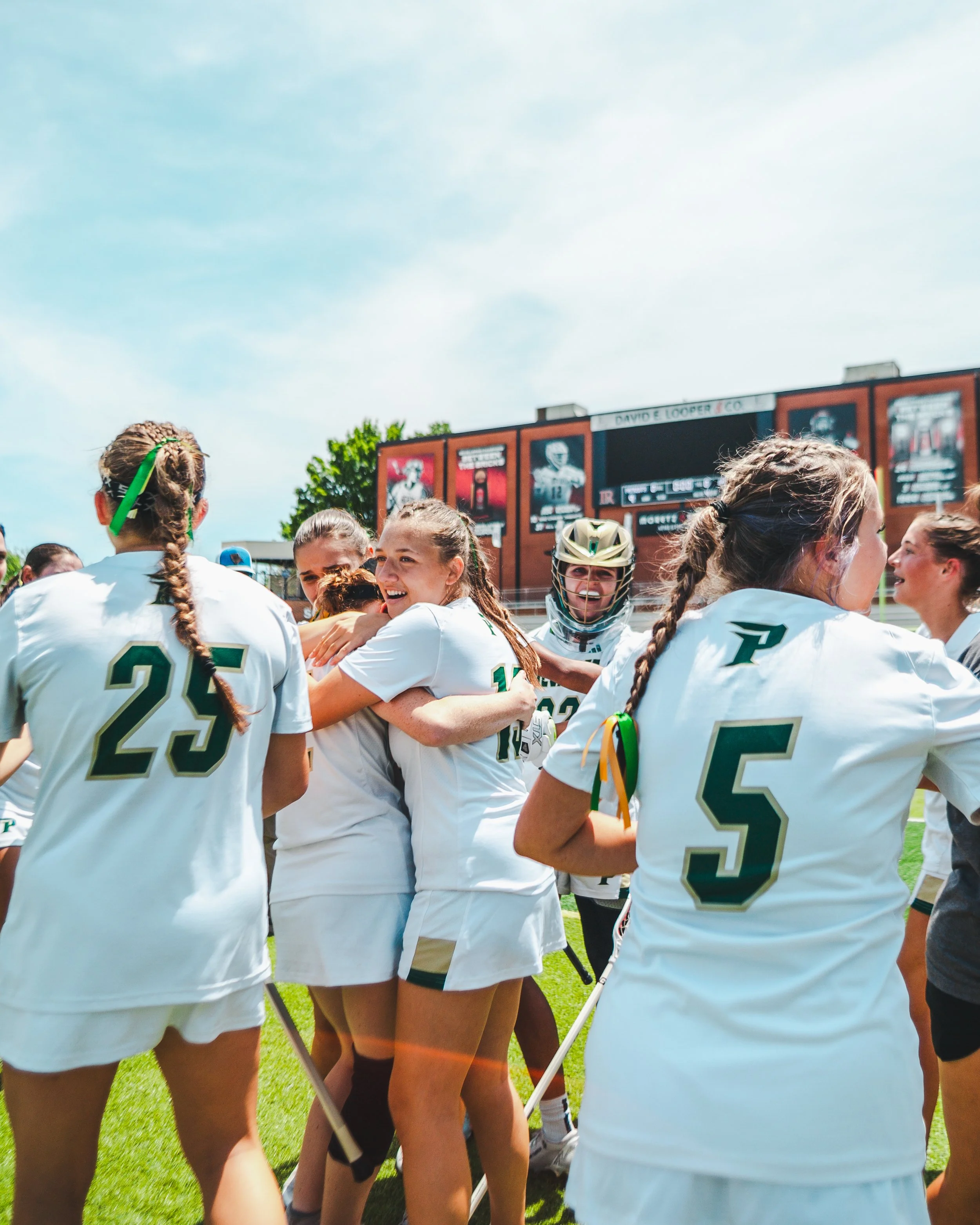 A group of girls in white sports uniforms gathers and celebrates on a bright green field during daytime.