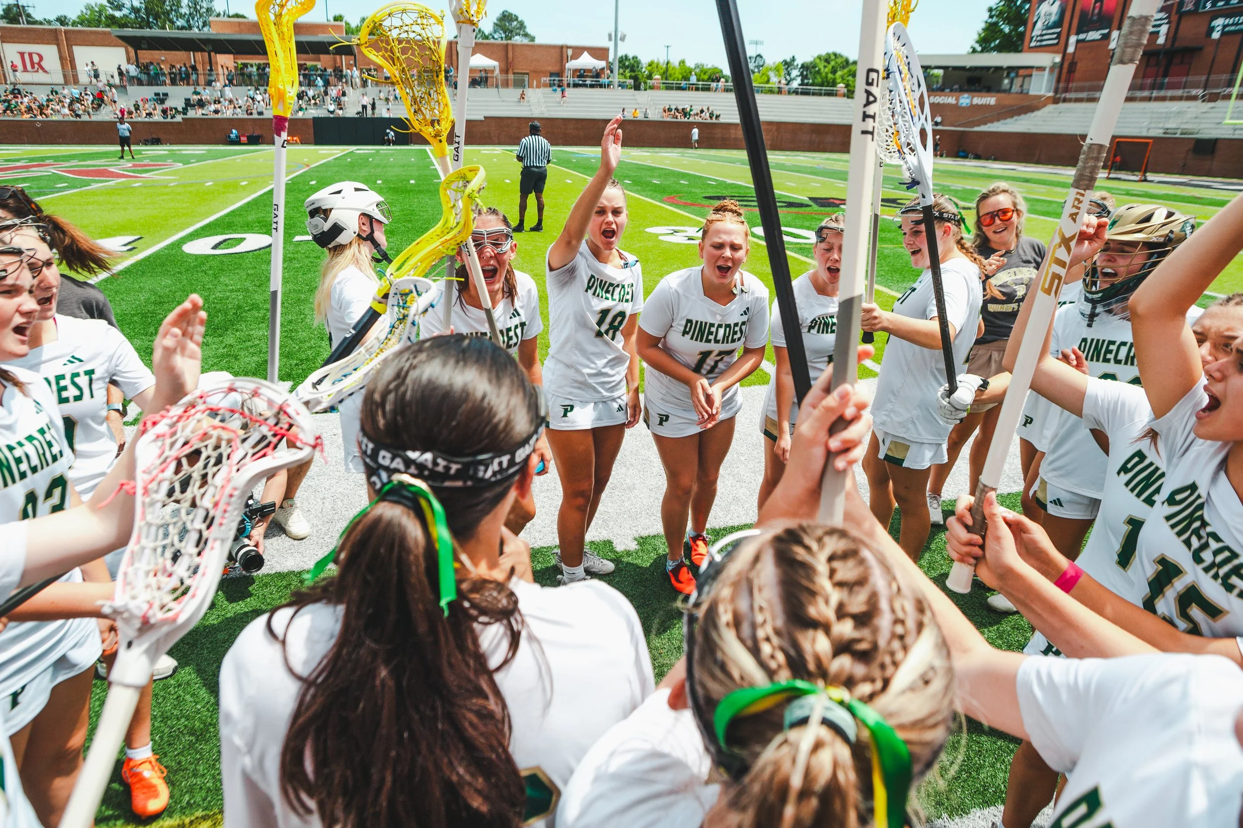 A girls lacrosse team celebrating on the field, some players with lacrosse sticks, in front of spectators in the stands.