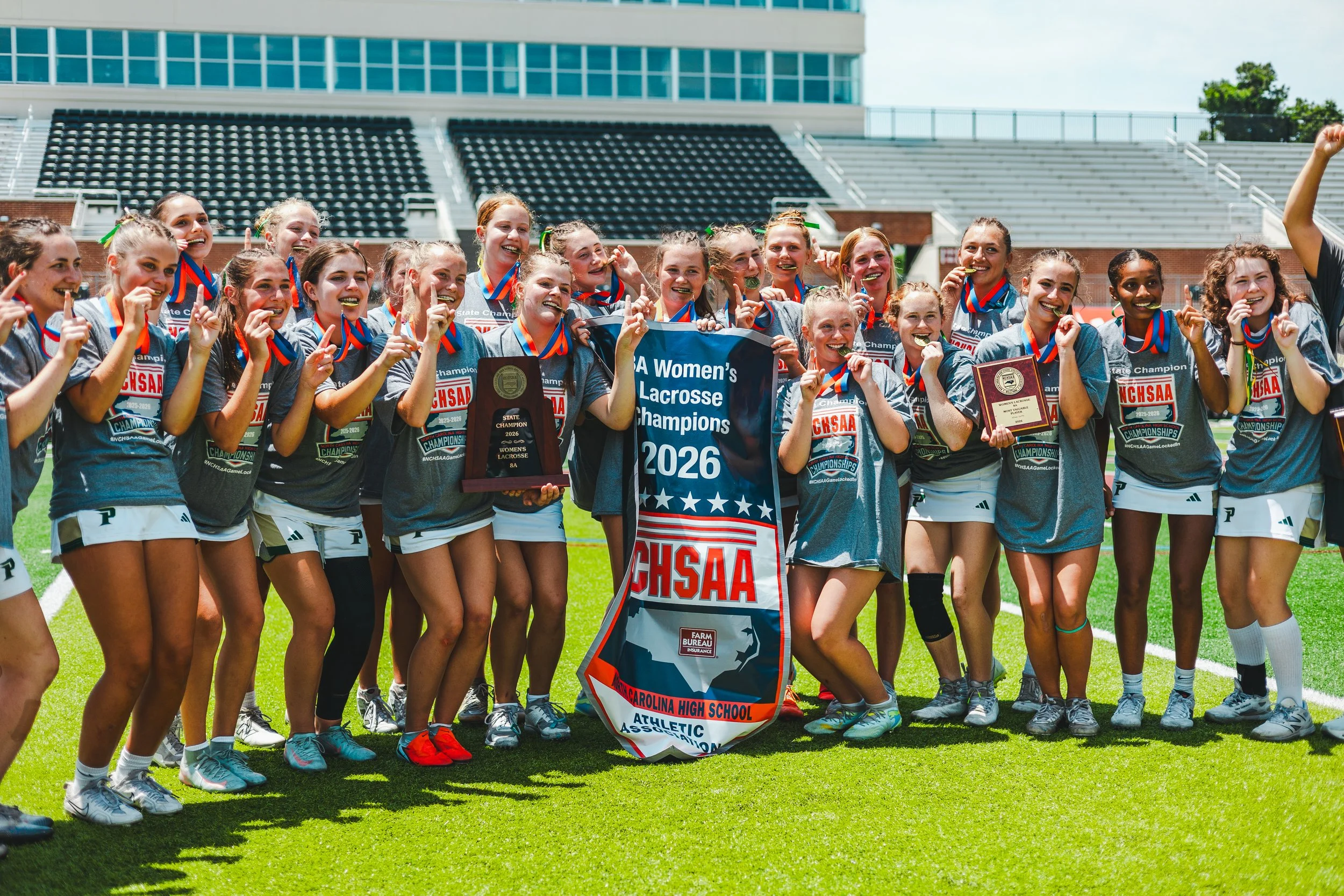 Group of young women lacrosse players celebrating on a field, holding medals, trophies, and a banner that reads "A Women’s Lacrosse Champions 2026," wearing team uniforms and smiling.