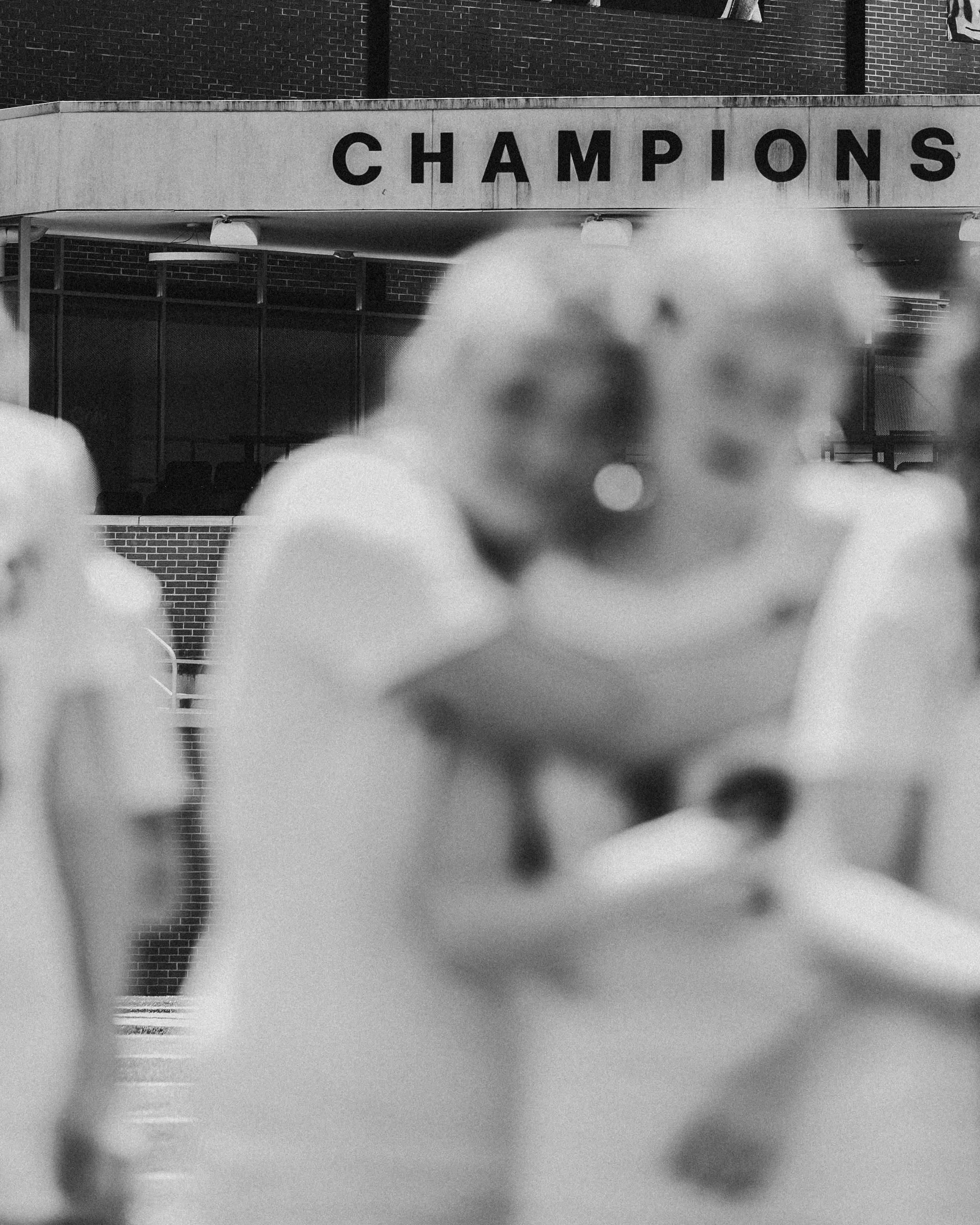 A black and white photo capturing a street scene with blurred objects in the foreground. In the background, there is a building with a sign displaying the word 'CHAMPIONS' on a concrete facade.