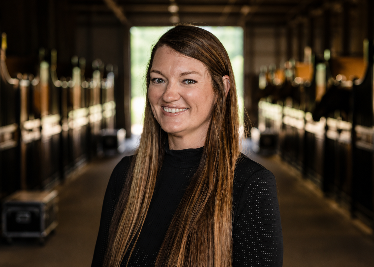 A woman with long brown hair smiling, standing inside a horse stable with wooden stalls and horses in the background.