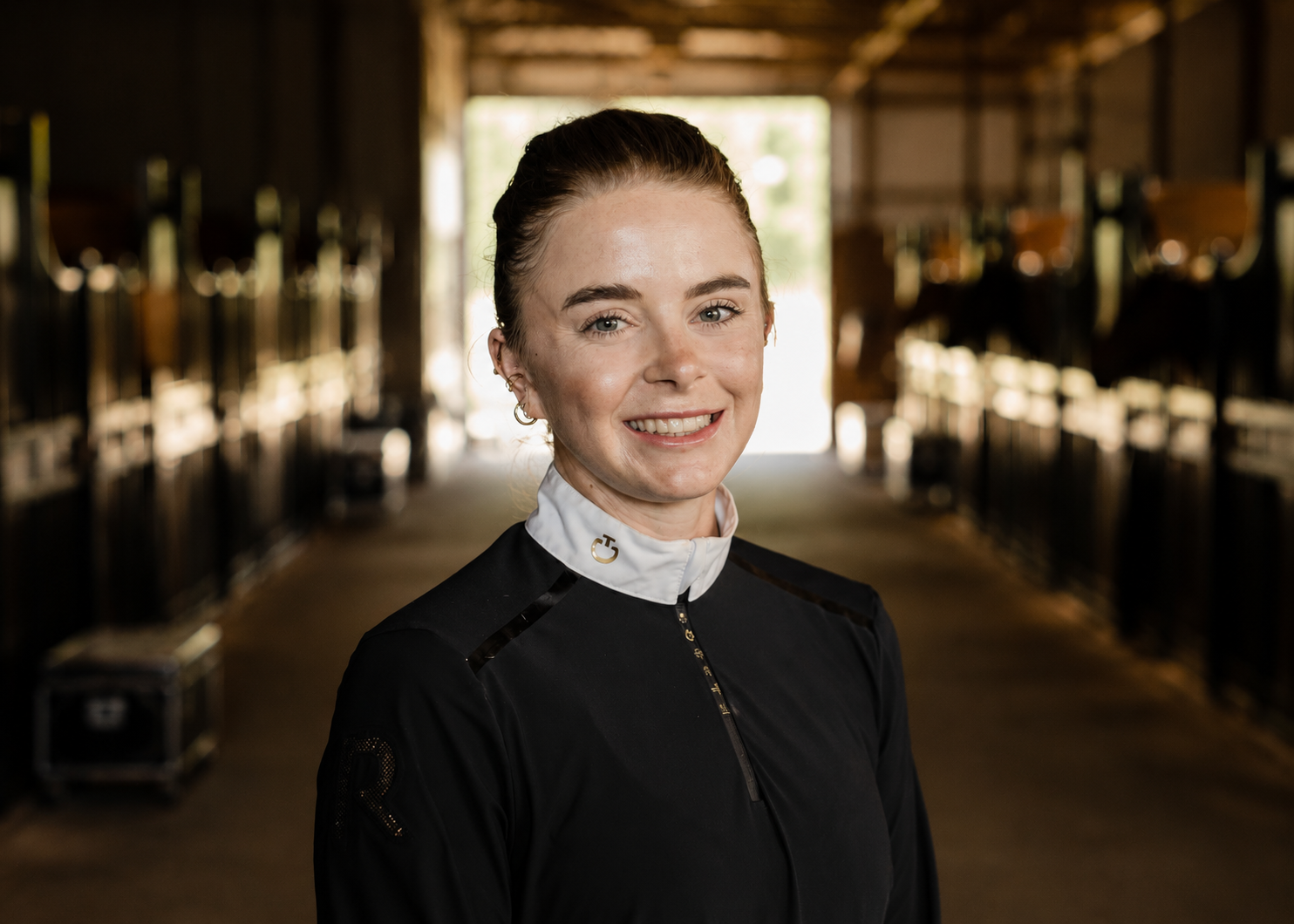 A smiling woman in a black athletic jacket with a white high collar stands inside a stable corridor, with horse stalls on both sides, illuminated by natural light from the open end in the background.