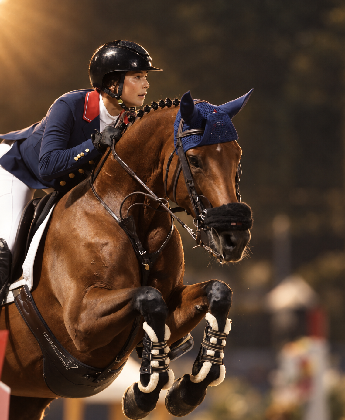 A female equestrian rider wearing a navy show jacket, white shirt, riding gloves, and a black helmet, riding a brown horse with a blue ear bonnet during a competition at sunset or evening.