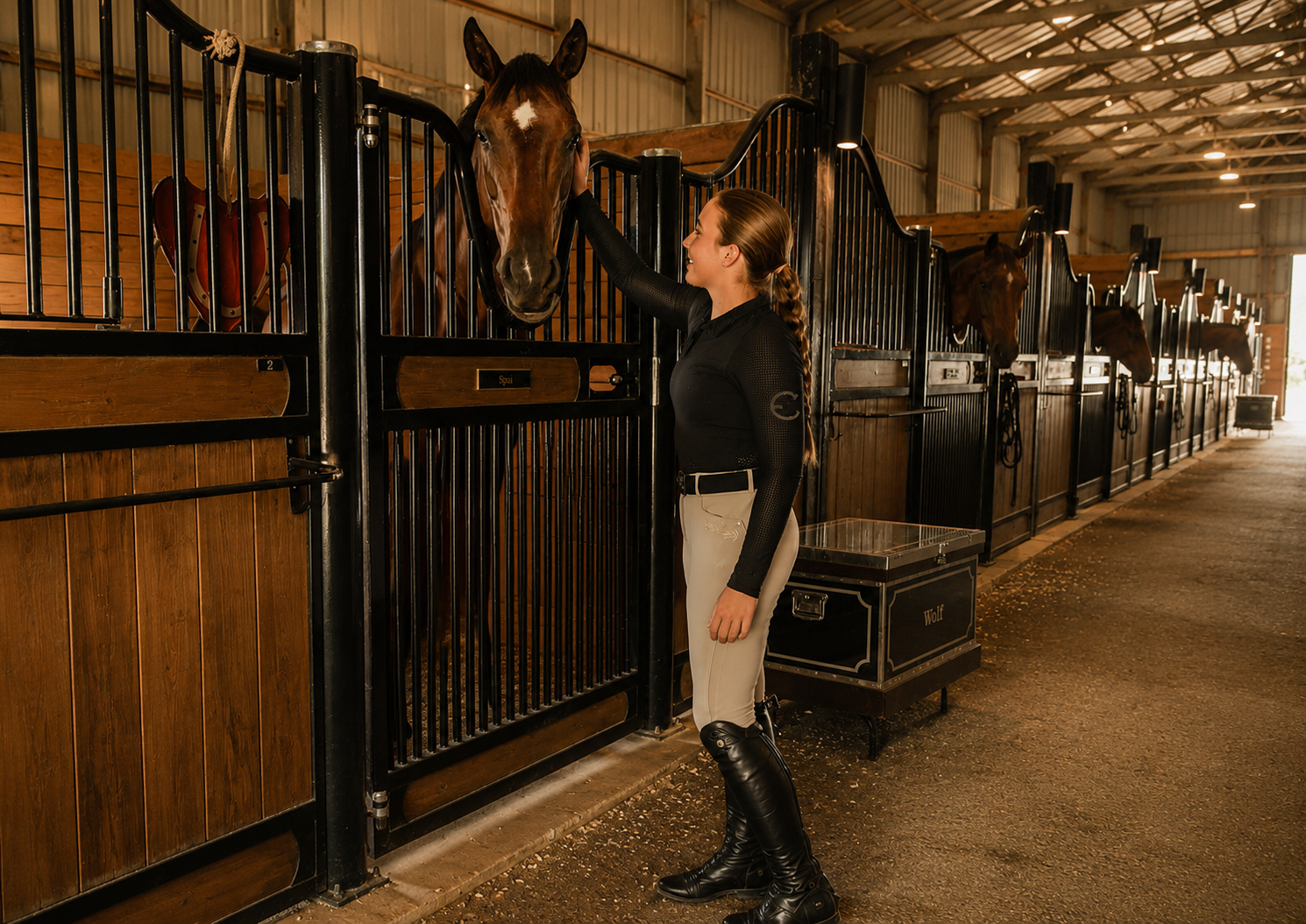 A woman grooming a brown horse in a stable with multiple horse stalls.