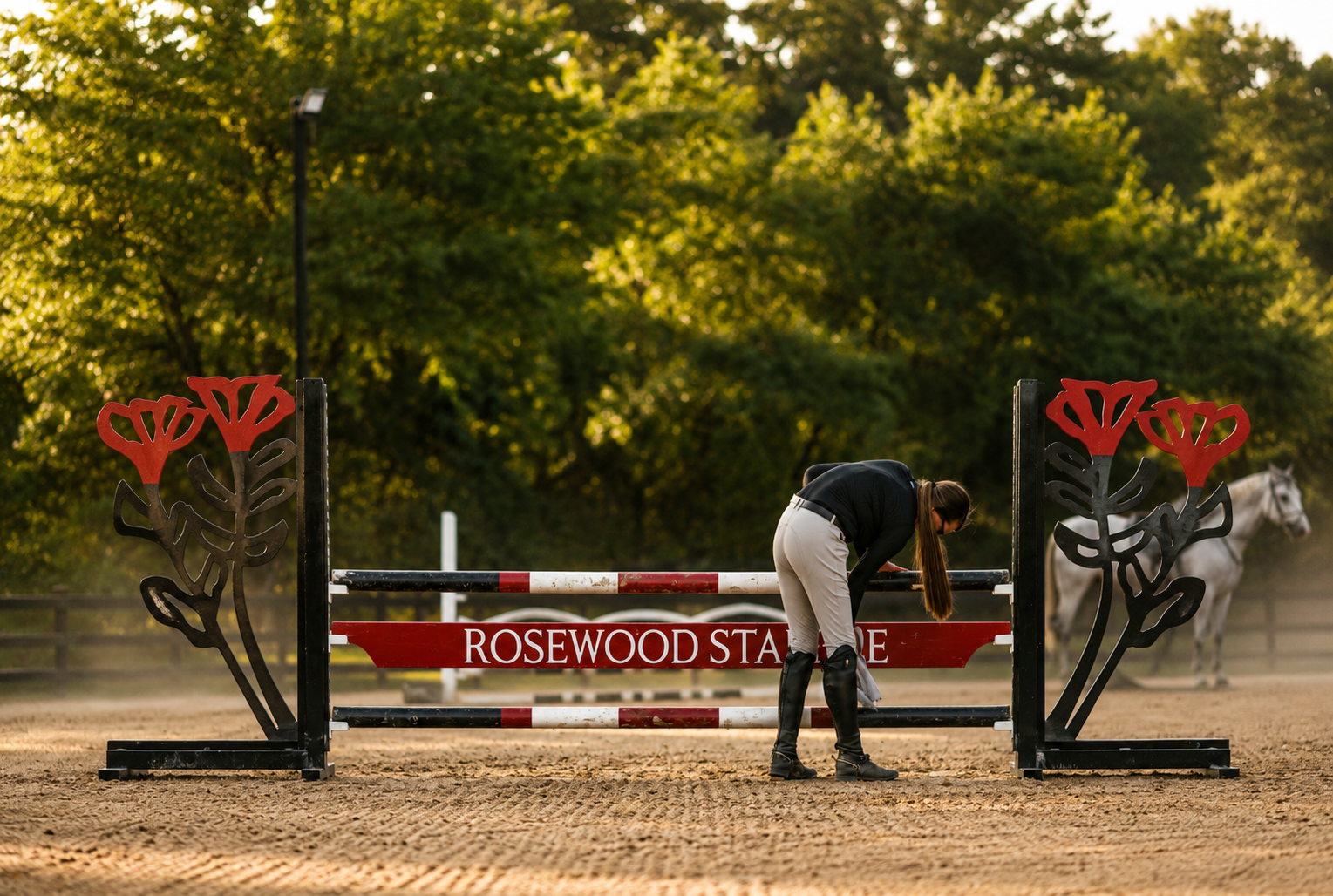 A woman adjusting a jump obstacle at Rosewood State Equestrian Center, with a white horse in the background on a dirt arena during daytime.