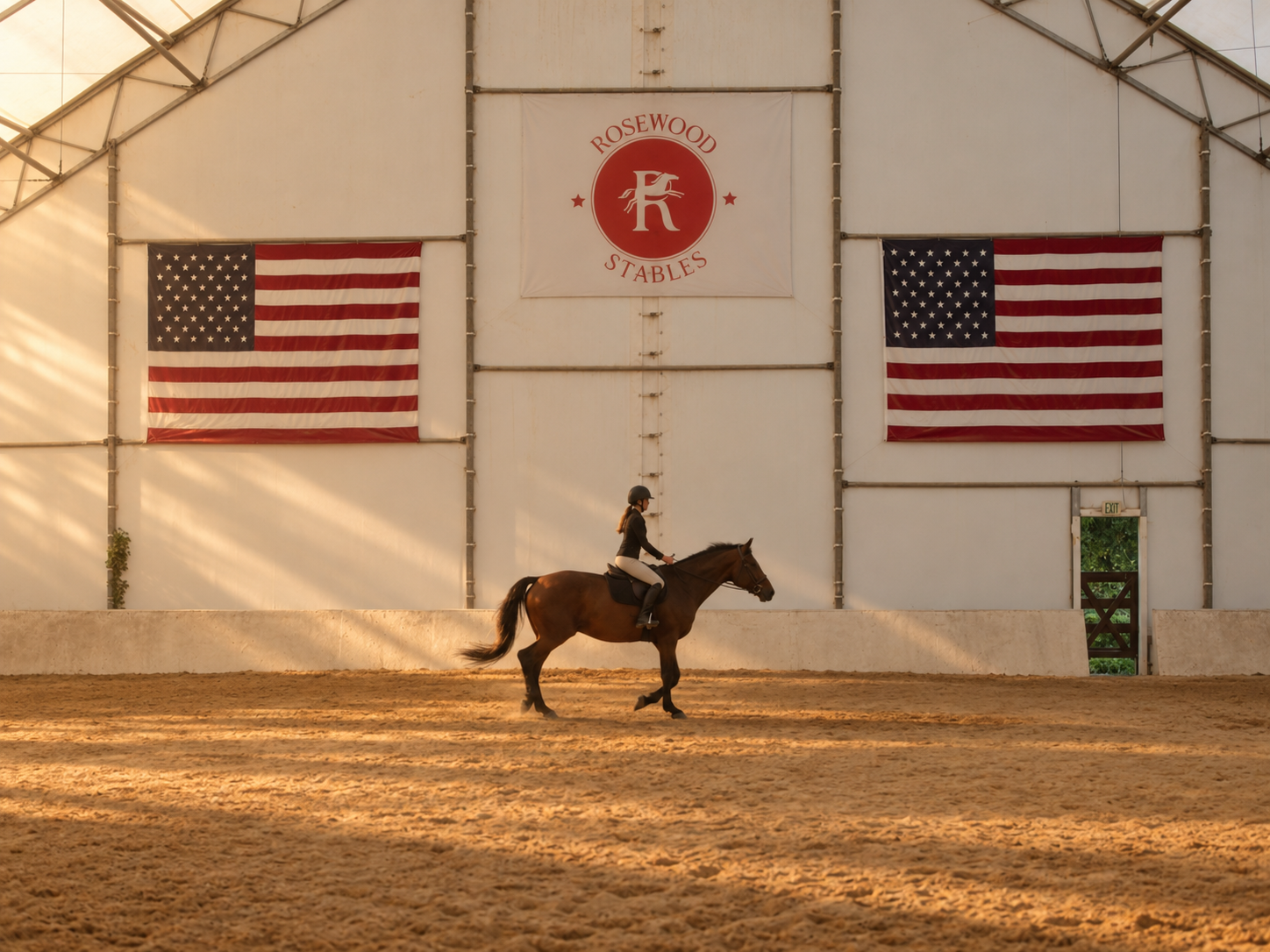 A person riding a horse inside an indoor riding arena, with American flags and a 'Rosewood Stables' banner on the wall.