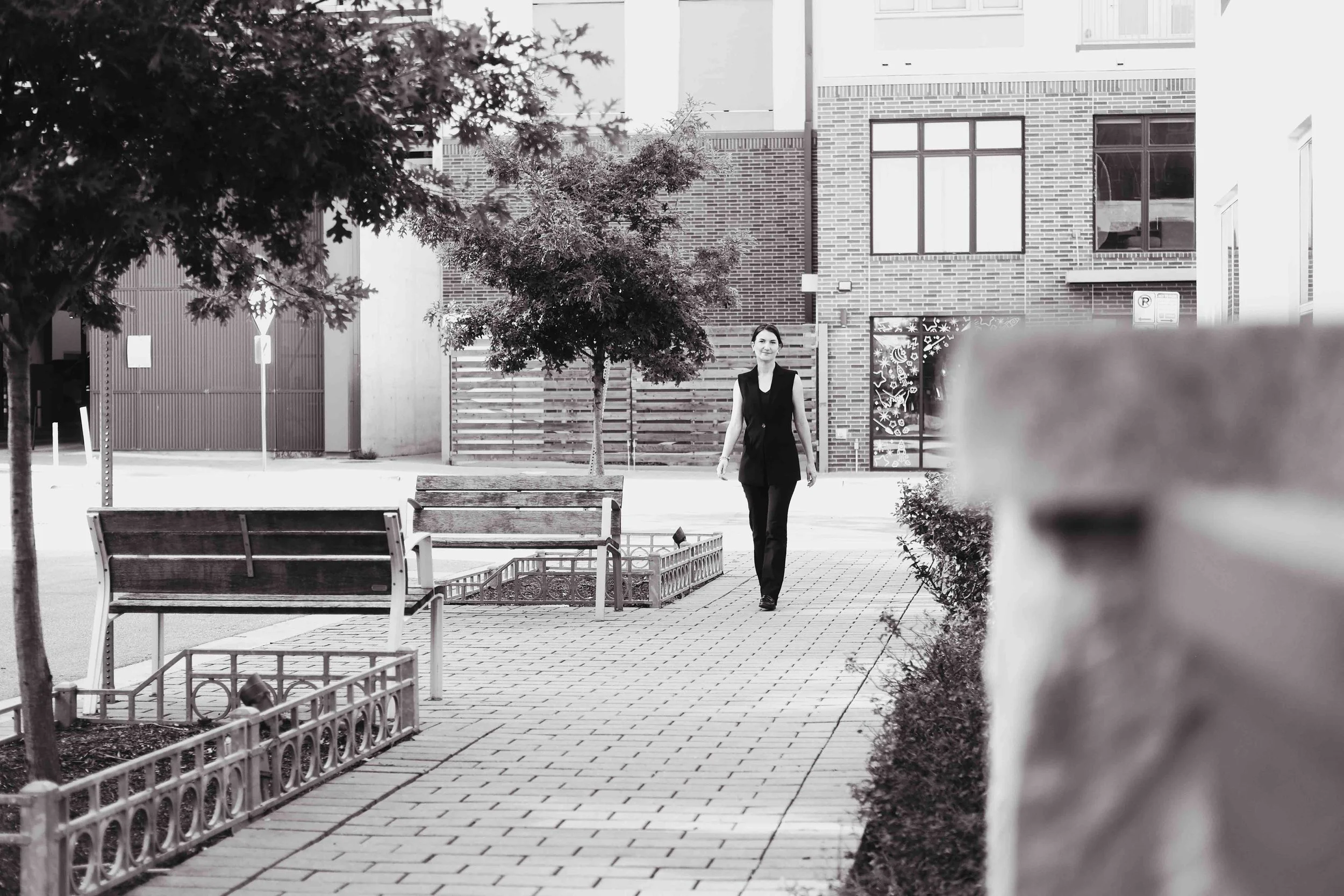 Black and white photo of Maureen Brown, M&A advisor, walking along a tree-lined brick path past park benches