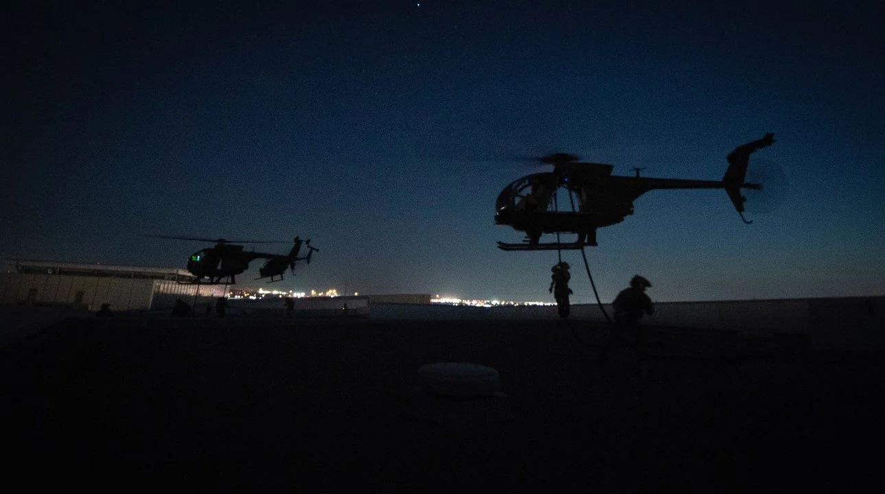 Two military helicopters on a rooftop at night with personnel working on one of the helicopters.
