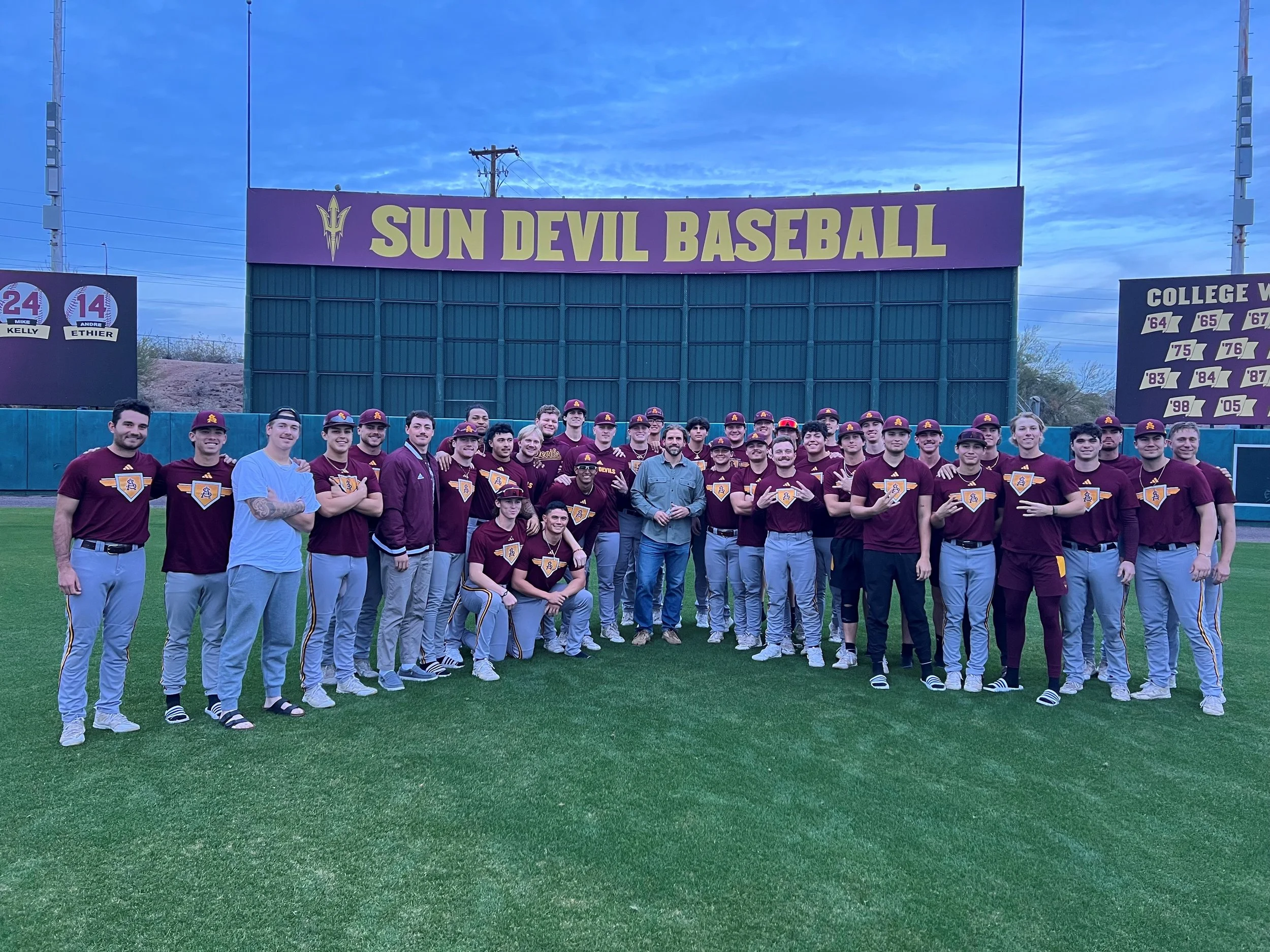 A group of baseball players and coaches standing on a baseball field in front of a large sign that says 'Sun Devil Baseball.' The players are wearing maroon uniforms with yellow and white accents, some have caps, and some are holding their gloves or trophies. The goal post and scoreboard are visible in the background amid a blue sky with some clouds.