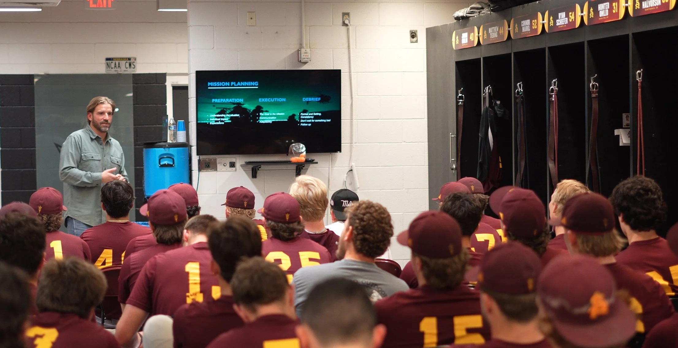 A man is giving a presentation to a group of students dressed in maroon caps and shirts with yellow numbers. The presentation slide on the screen reads 'Mission Planning' with sections 'Preparation,' 'Execution,' and 'Debrief.' The setting appears to be a locker room with lockers and sports gear.