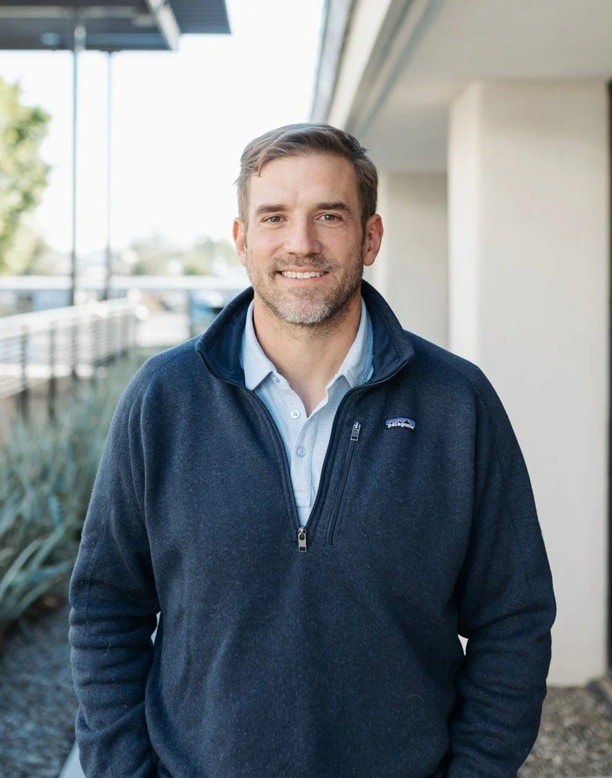 A smiling man with light brown hair and a beard, wearing a navy Patagonia fleece over a light blue collared shirt, standing outdoors near a building with a modern architectural style.