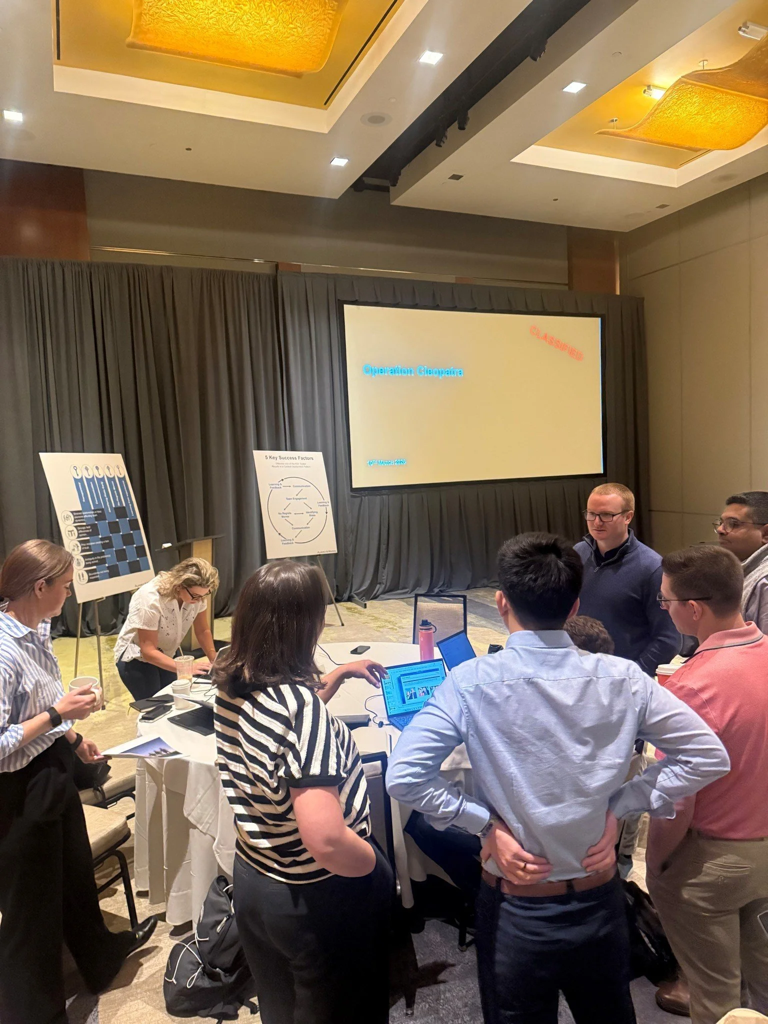 A team of professionals gathered around a table works through a mission-planning exercise during a multi-day leadership program, applying communication, decision-making, contingency planning, and teamwork to a complex scenario.