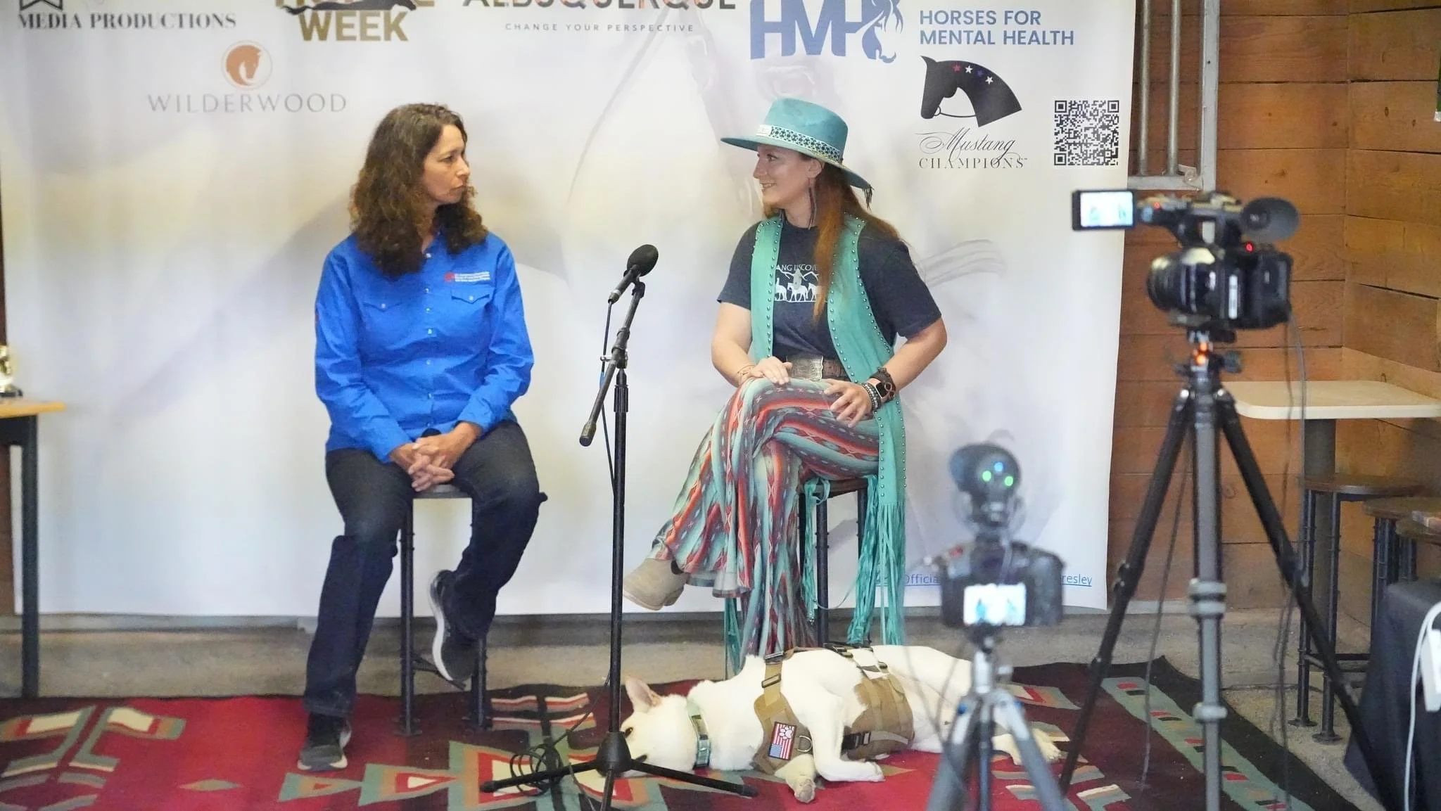 Two women sitting in front of a backdrop with logos and banners, one wearing a blue shirt and the other wearing a hat and colorful skirt, during a recorded interview or discussion. There are cameras and microphones capturing the scene.