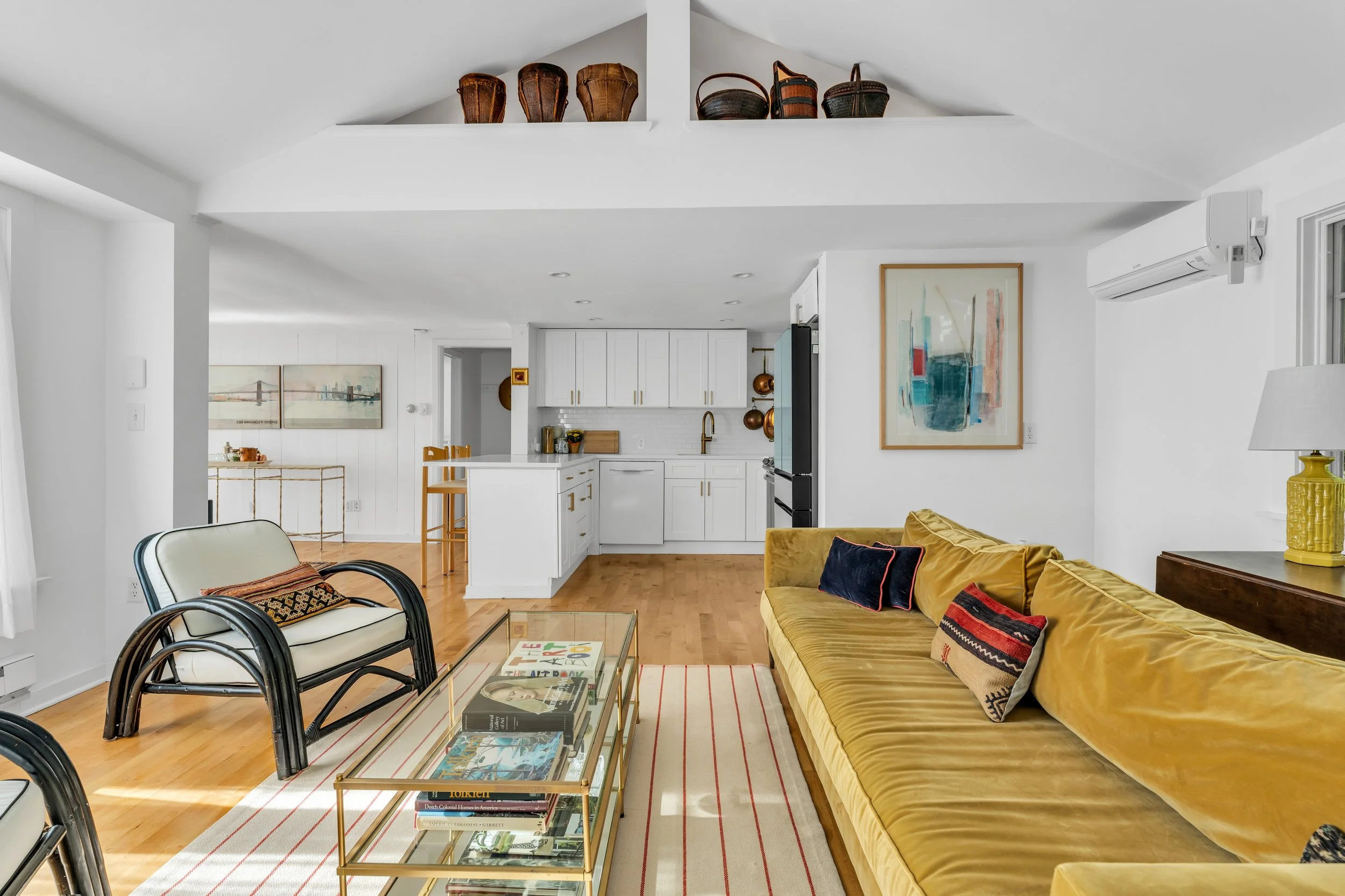 A living room with white walls and ceiling, featuring a mustard yellow velvet sofa with decorative pillows, a glass coffee table with magazines, a white armchair with a colorful pillow, and wall art including a framed abstract painting. The room opens into a kitchen with white cabinetry, brass hardware, and wooden accents, and has a wooden floor and minimal decor.