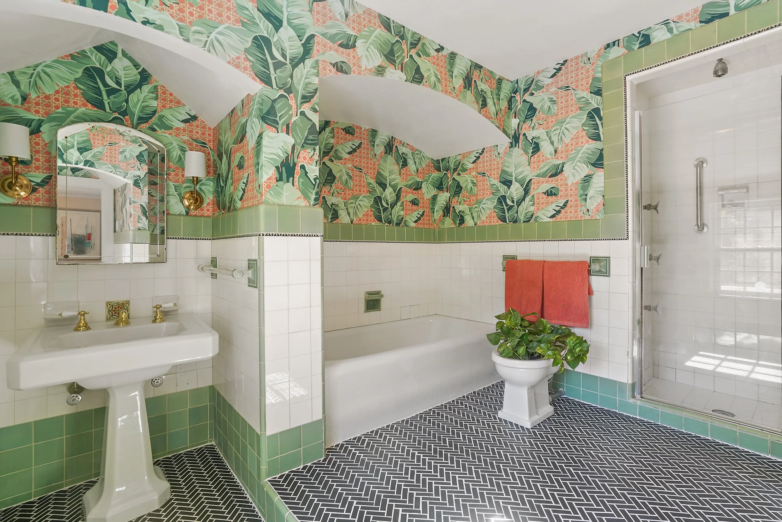 Bathroom with tropical leaf wallpaper, green tile accents, black and white herringbone floor, white sink with gold fixtures, mirror, orange towel, and plants.
