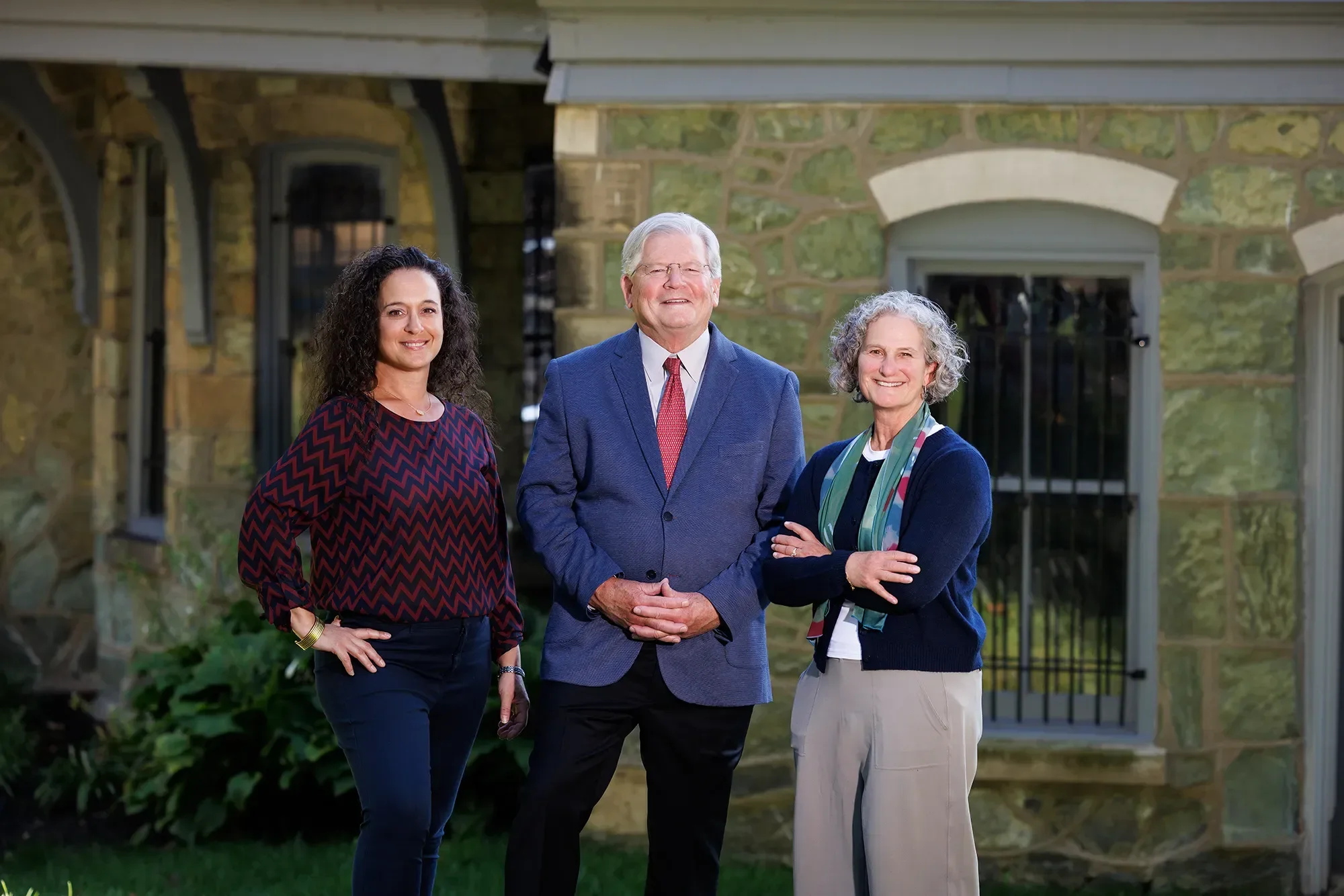 Mariana, Jim, and Amy standing outdoors in front of a stone building with arched windows and a gate, smiling at the camera.
