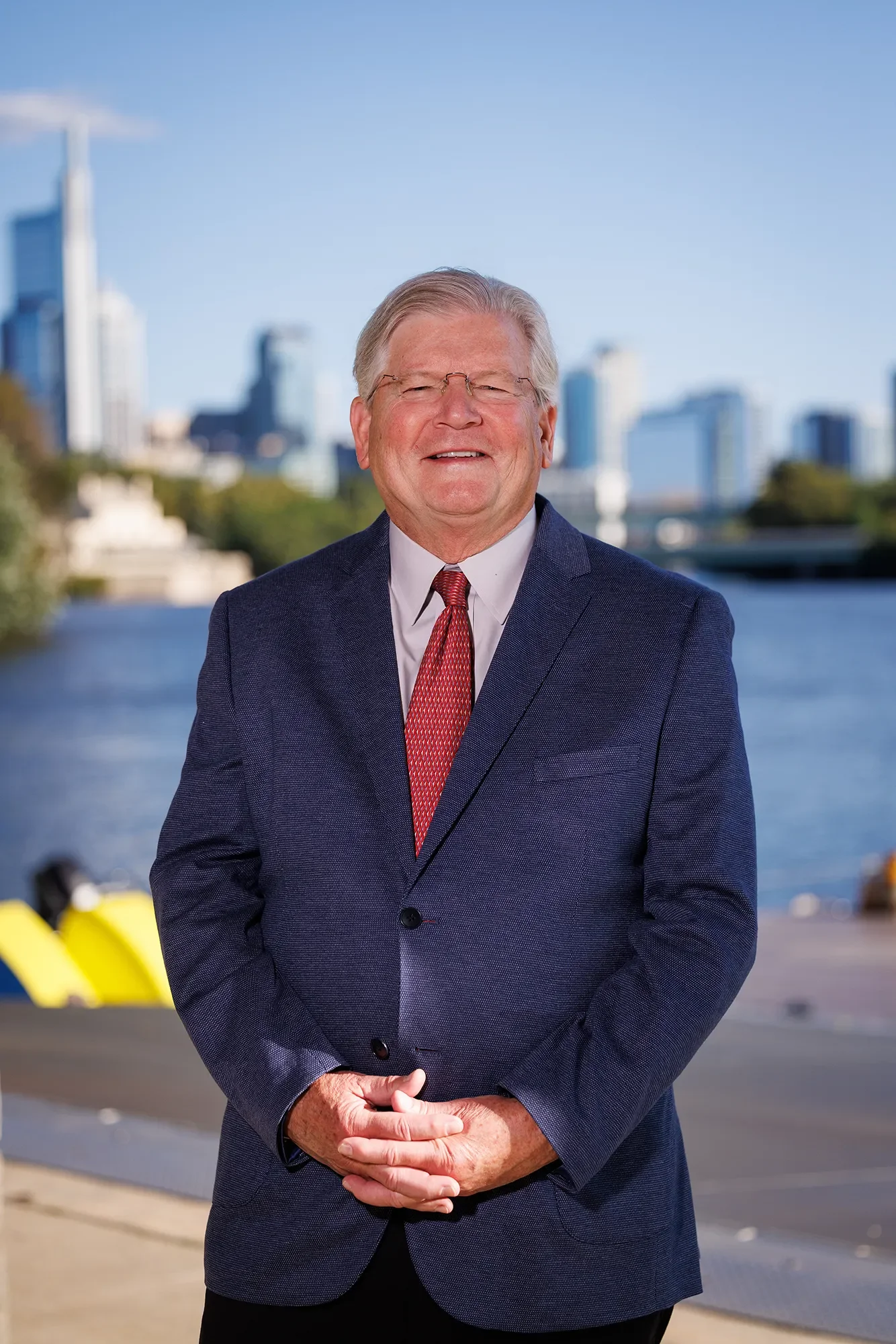 Jim Rohn in a navy blue suit with a red tie, standing in front of the Philadelphia skyline, smiling at the camera.