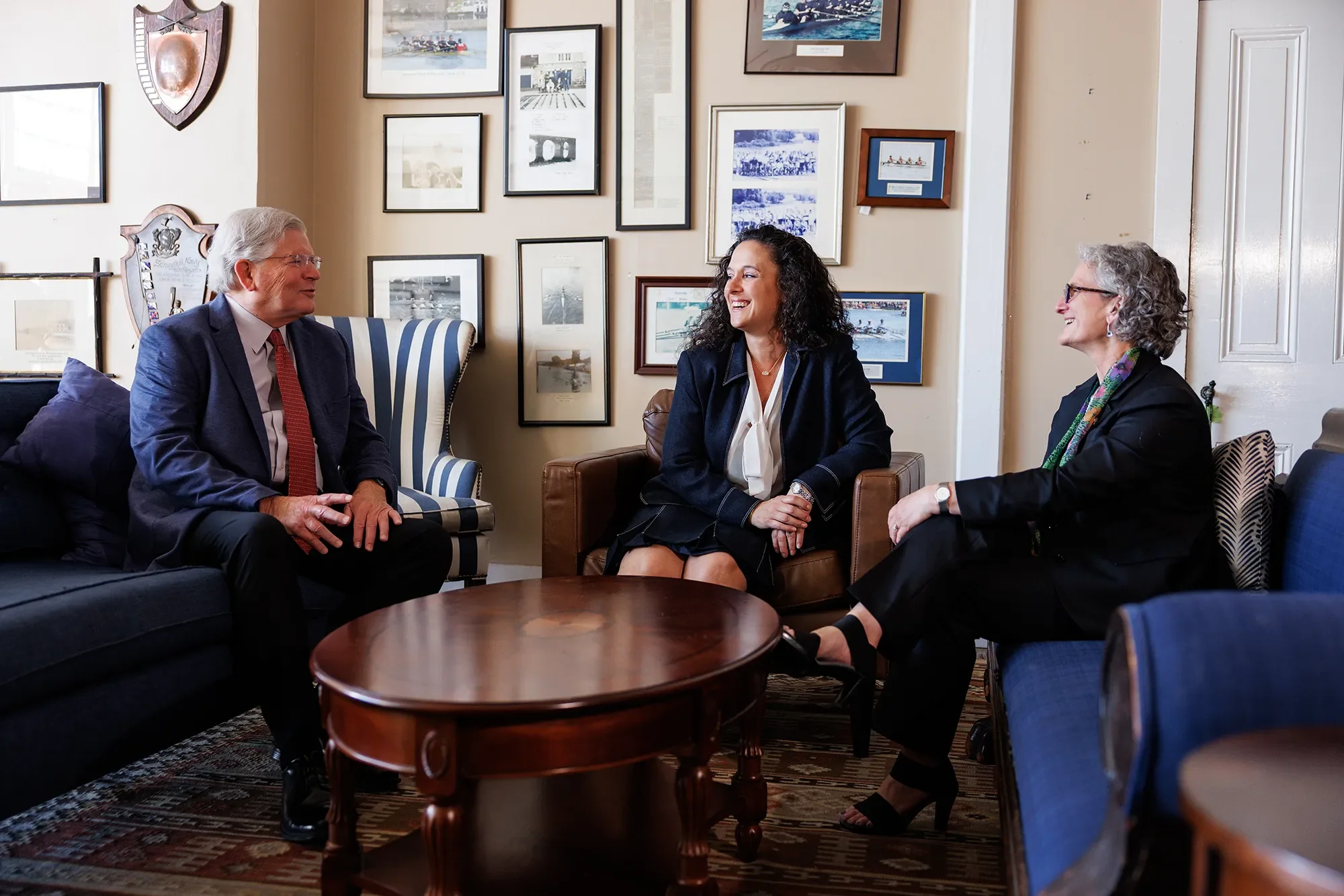 Jim, Mariana, and Amy, seated and talking in a room decorated with various framed photographs.