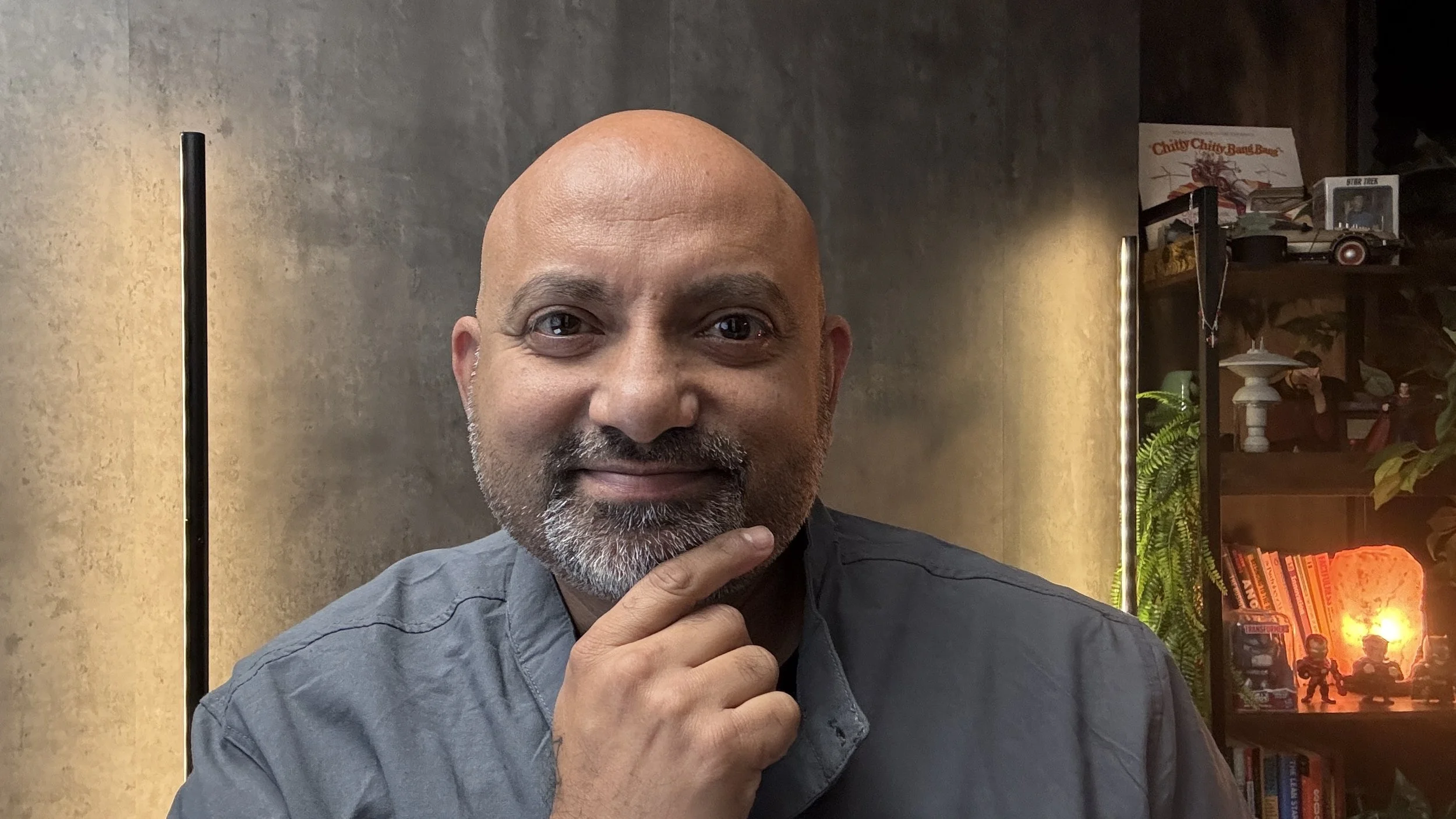Ajay Chikermane, founder of NowHuman with a bald head and gray beard smiling while touching his chin, indoors with a bookshelf and decorative items in the background.