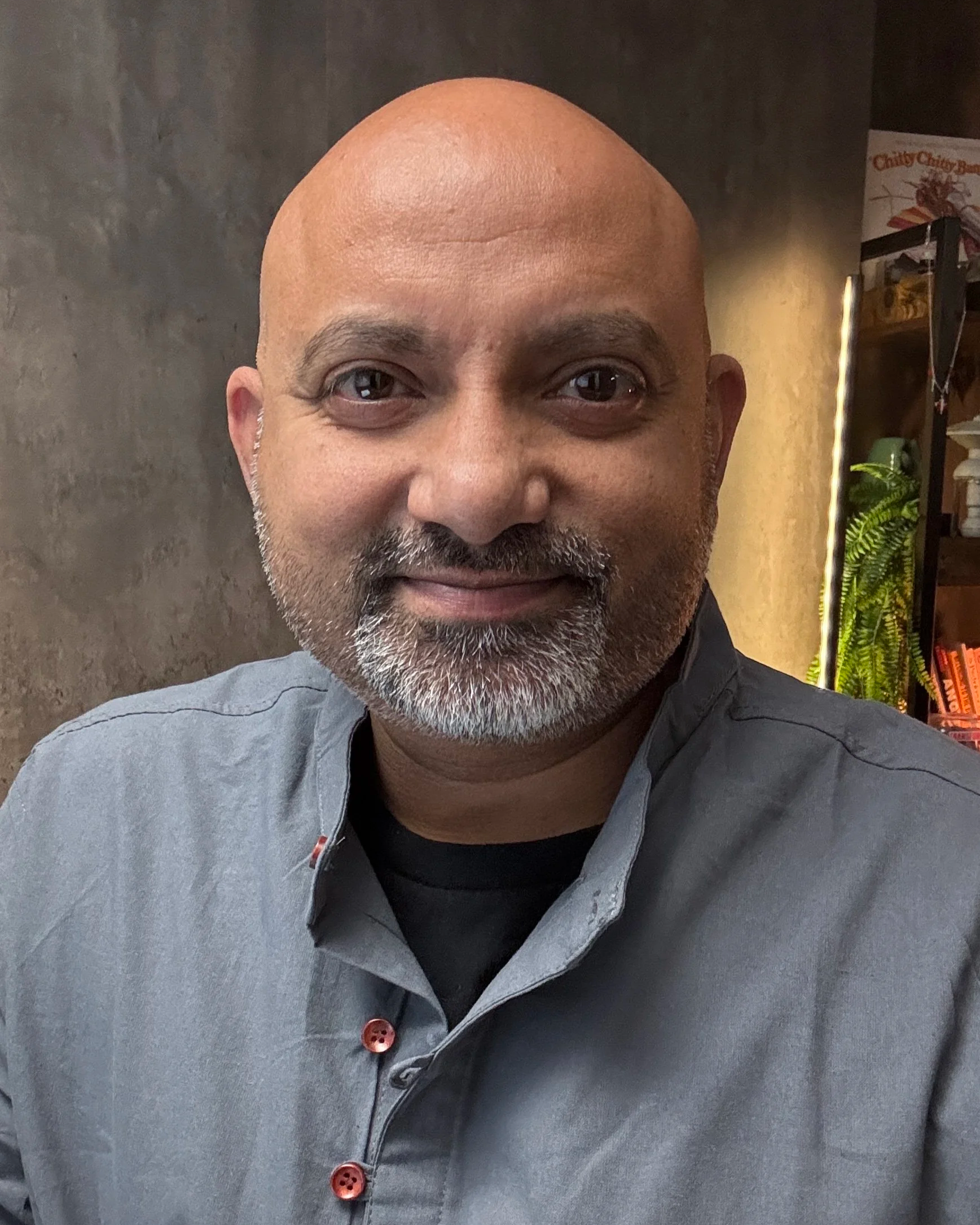A close-up of Ajay Chikermane's face, showing his gray beard, bald head, and brown eyes. The background displays a dark wall and part of a bookshelf with books and a plant.