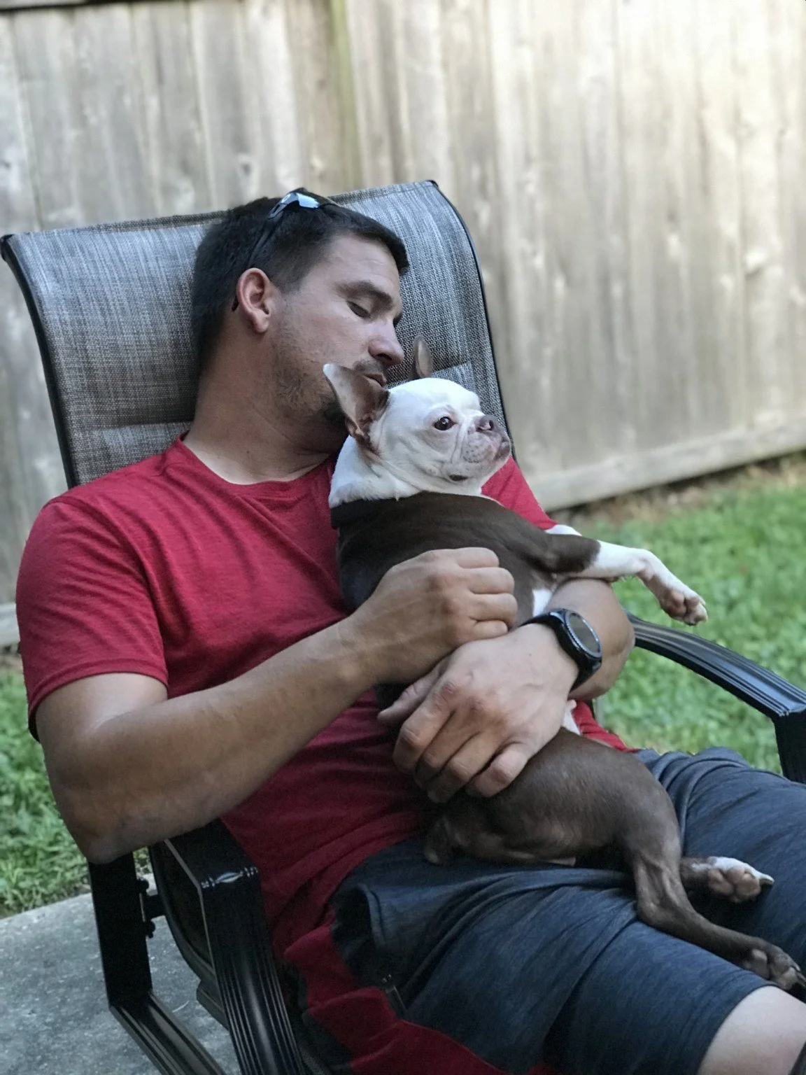 Man sitting outdoors holding a Boston Terrier on his lap.