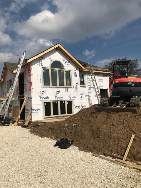 Construction of a house with exterior walls covered in Tyvek house wrap, showing large arched and rectangular windows, construction equipment, and ladders on a partly cloudy day.