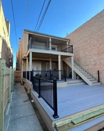 Newly built two-story wooden house with a deck on both levels, black metal railings, and exterior stairs leading to the upper deck, located between two brick buildings under a clear blue sky.