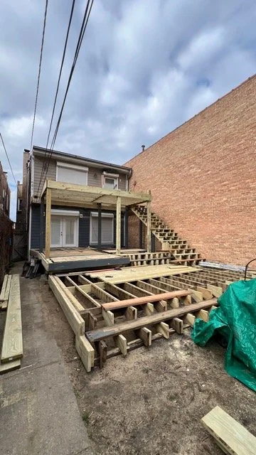 Construction site with a wooden deck being built in the backyard, with stairs leading up to the deck. The house has a second-floor balcony. The sky is partly cloudy.