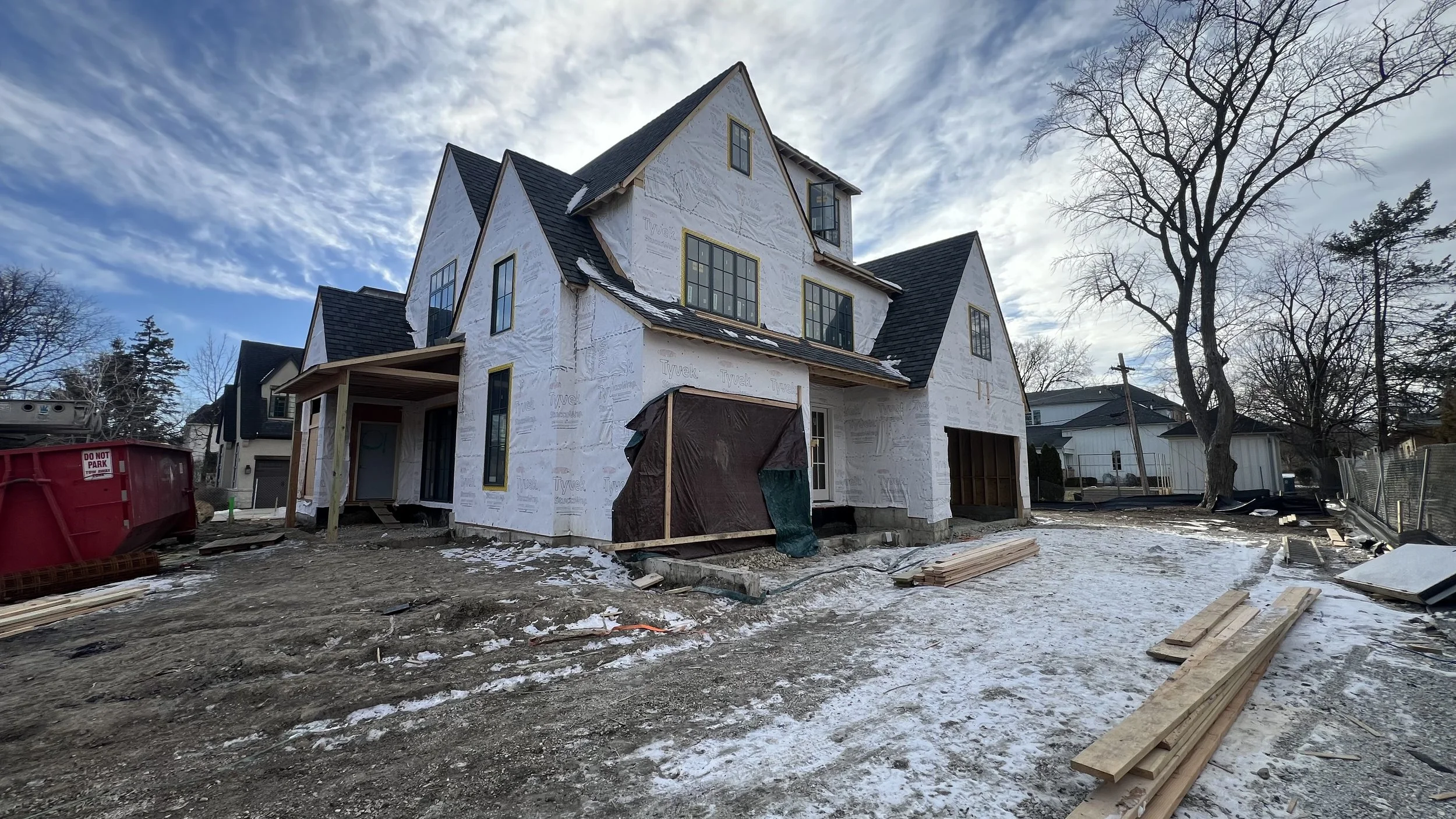 A house under construction with exterior walls covered in white sheathing, multiple large windows, and a steep gabled roof. Construction materials and debris are scattered on the ground, with some snow and dirt visible. A leafless tree stands nearby 