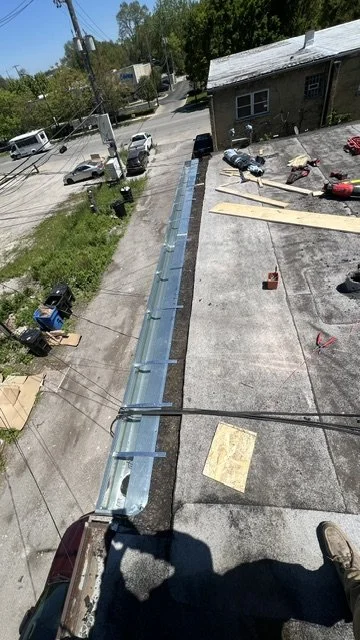 View from a rooftop showing a construction project on the roof with tools, wood, and equipment, adjacent to a residential area with streets, cars, and trees.