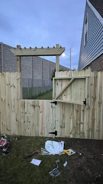 View of a backyard wooden fence with a gate under construction, tools and materials on the ground, a wooden frame for a garden arch or trellis in the background, and neighboring houses visible.