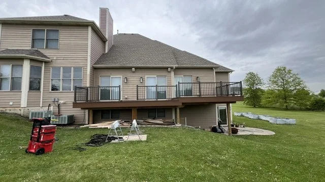 Backyard view of a house with a wooden deck, patio furniture, gardening tools, and a cloudy sky.