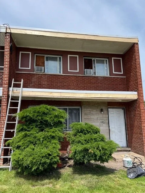 Two-story apartment building with brick exterior, balcony, and windows with air conditioners, garden bushes in front, ladder leaning against the wall, and household items near the entrance.