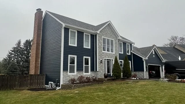 Modern two-story house with dark blue and gray siding, front lawn, and a garage with a vehicle parked outside.