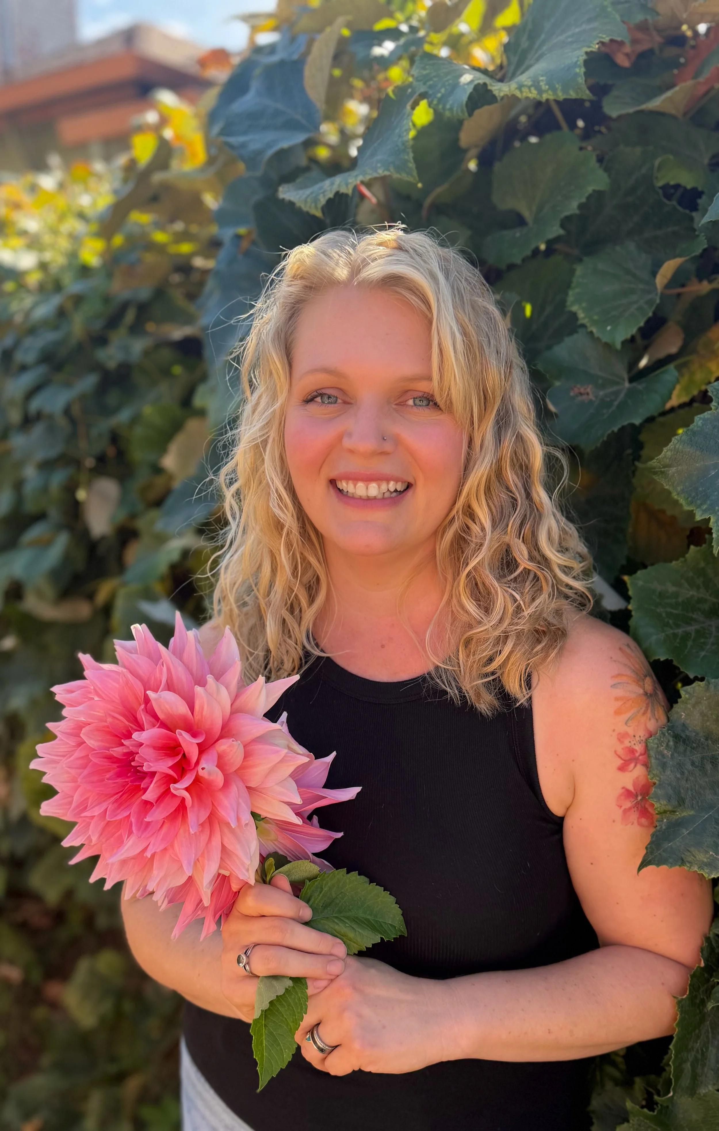 A woman with blonde curly hair, wearing a black sleeveless top, smiling, holding a large pink dahlia flower, standing in front of green leafy plants.