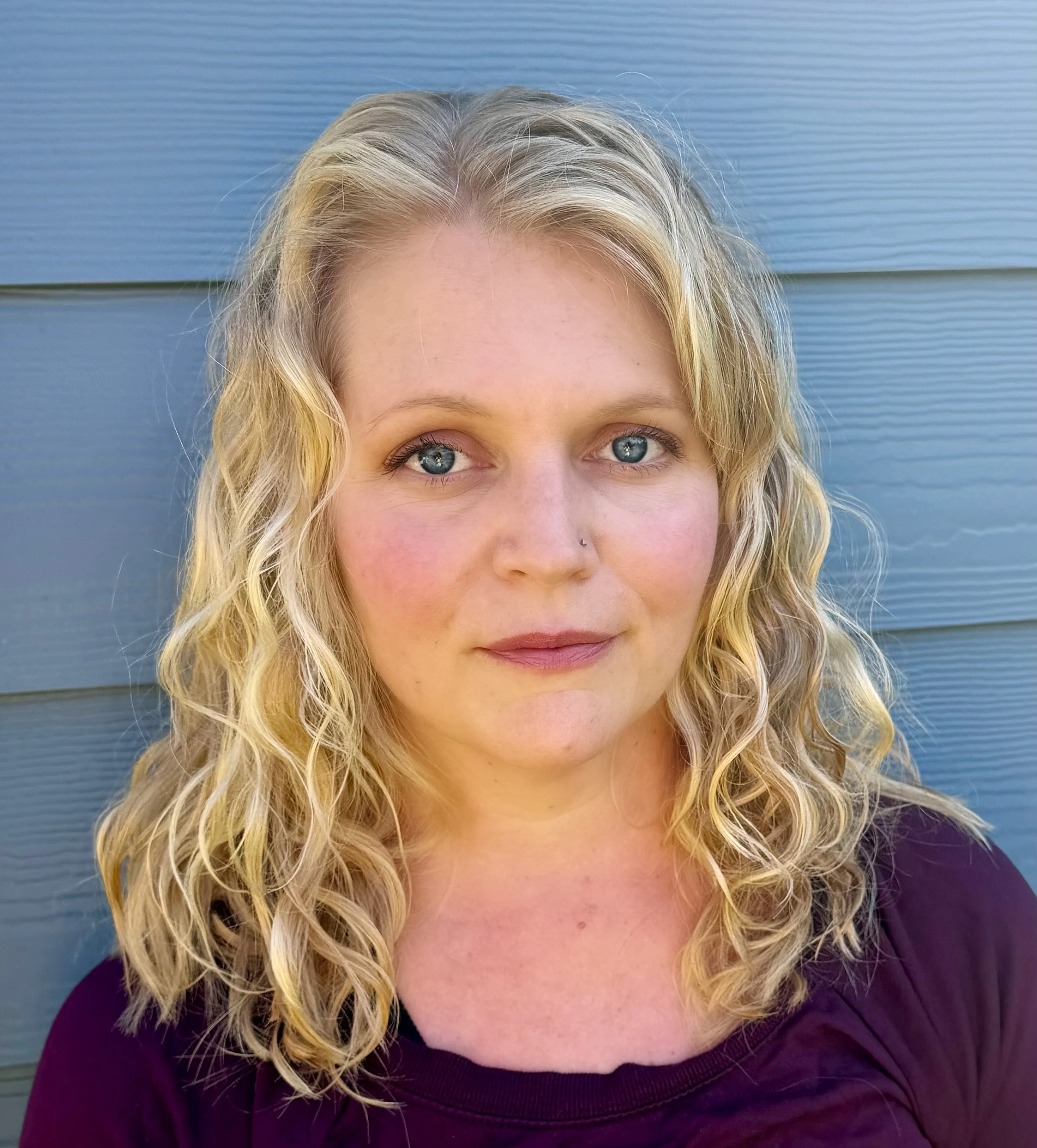 Close-up of a woman with blonde curly hair, blue eyes, and fair skin, standing in front of a blue wooden wall.