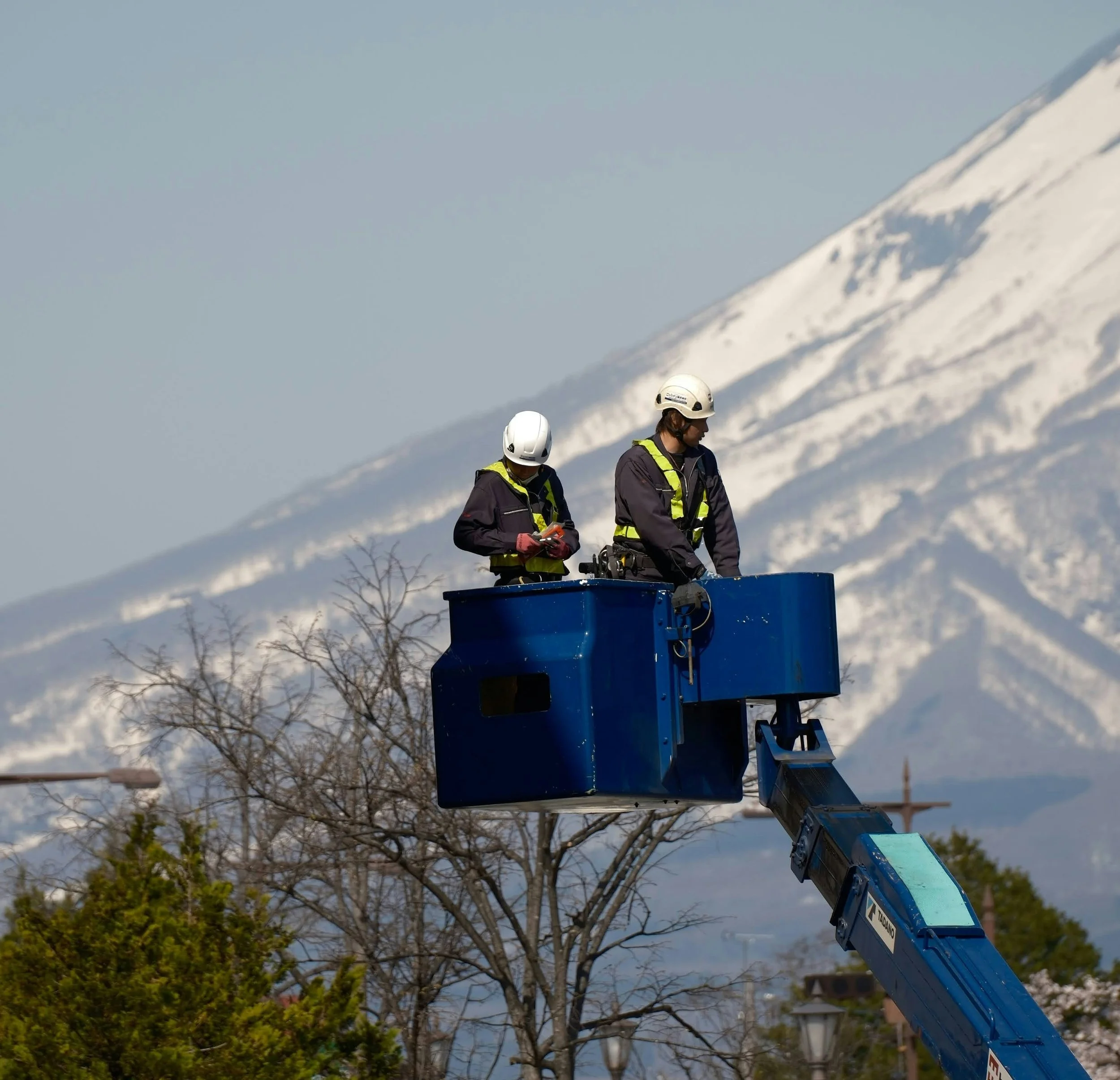 Two workers wearing helmets and reflective vests are standing in an elevated blue bucket lift, working on or inspecting something. There are trees and a snow-capped mountain in the background.