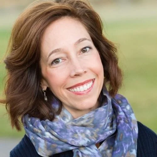 A woman with shoulder-length brown hair smiling outdoors, wearing a blue scarf and earrings.