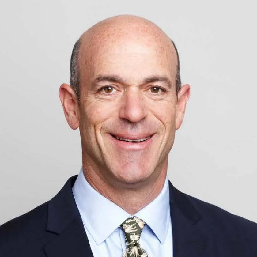 Headshot of a smiling middle-aged man with a bald head, wearing a dark suit, light blue shirt, and patterned tie against a neutral background.