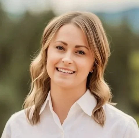 A young woman with blonde hair smiling outdoors, wearing a white shirt.