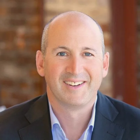 Headshot of a smiling man with blue eyes, and a shaved head, wearing a dark blazer and light blue shirt, with a brick wall background.
