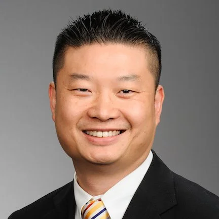 Headshot of a smiling man in a suit and striped tie against a gray background.