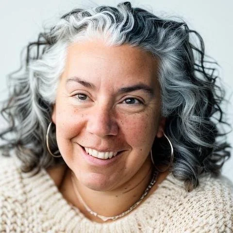 A woman with curly, gray and black hair smiling at the camera, wearing a beige knit sweater and hoop earrings.