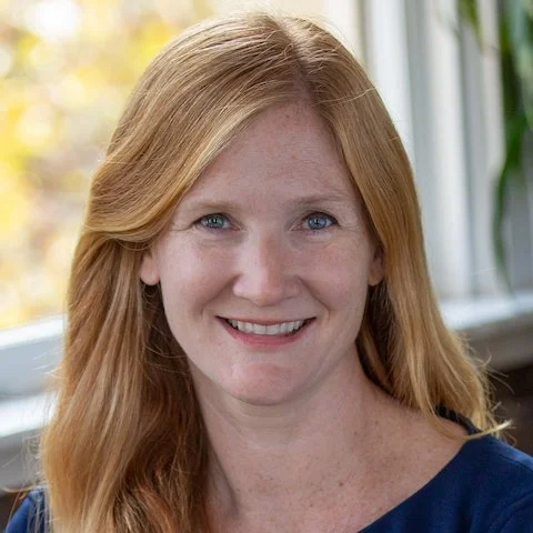 A woman with long red hair and blue eyes smiling at the camera, standing in front of a window with natural light.