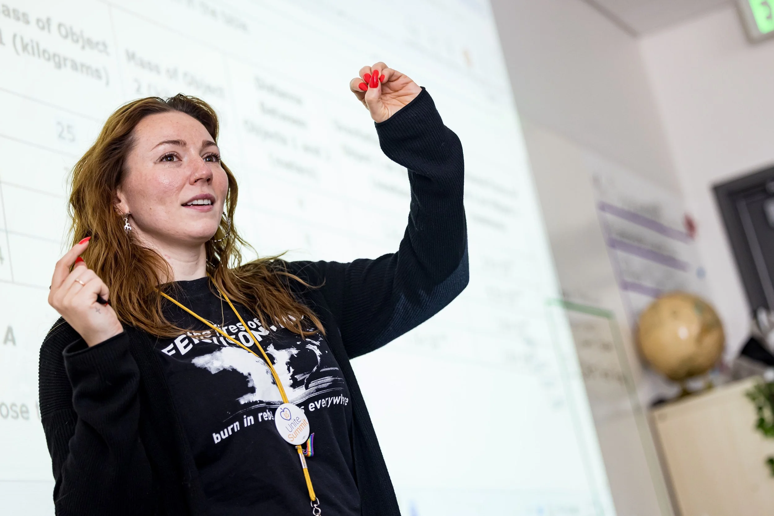 A woman with red hair and red lipstick is speaking or teaching in a classroom, gesturing with her right hand. She is wearing a black shirt with text and a button badge that says "Unite Summit". Behind her is a whiteboard with writing on it.