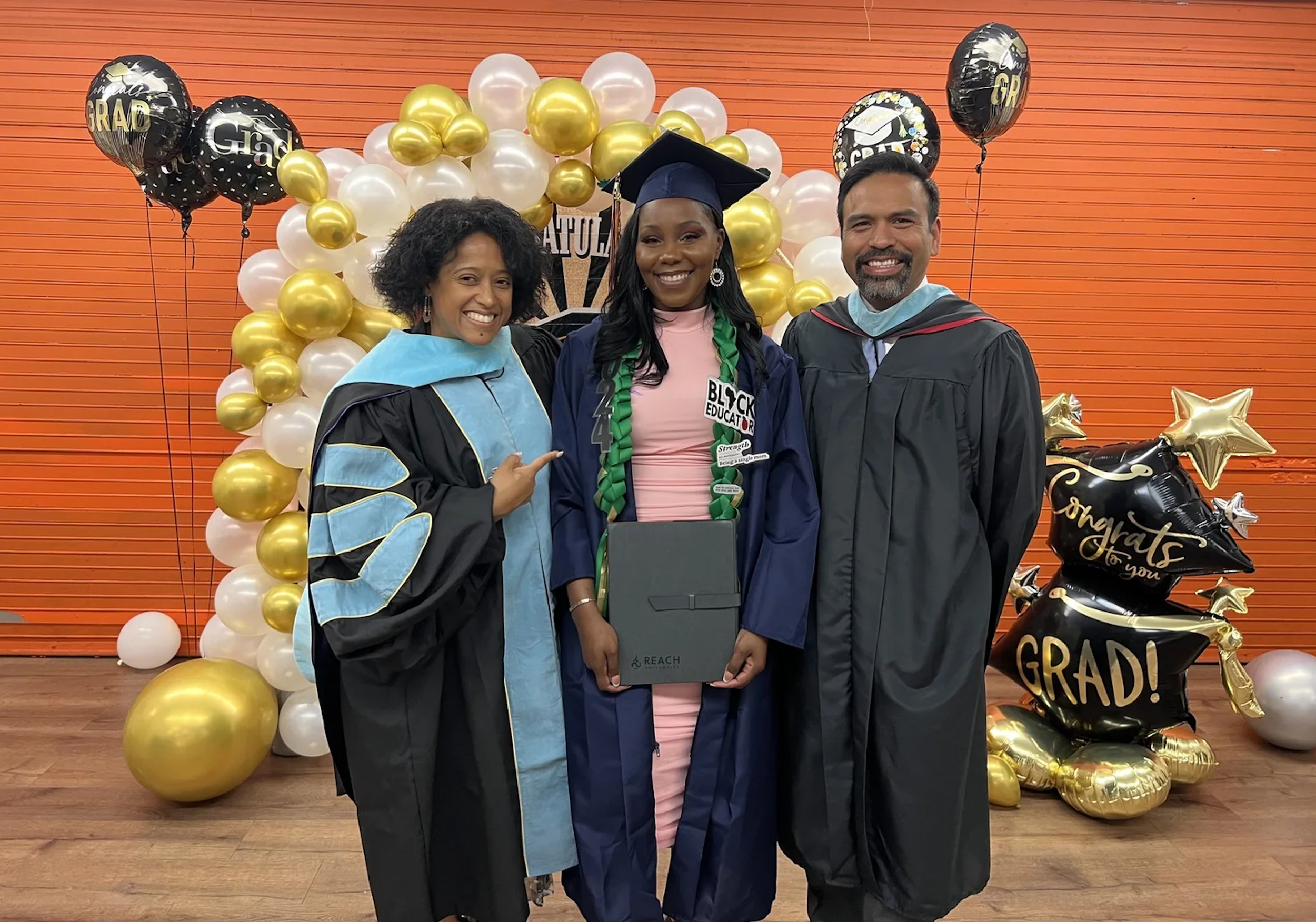 A woman in graduation attire standing between two adults at a graduation celebration, holding a diploma, with a balloon arch behind them.